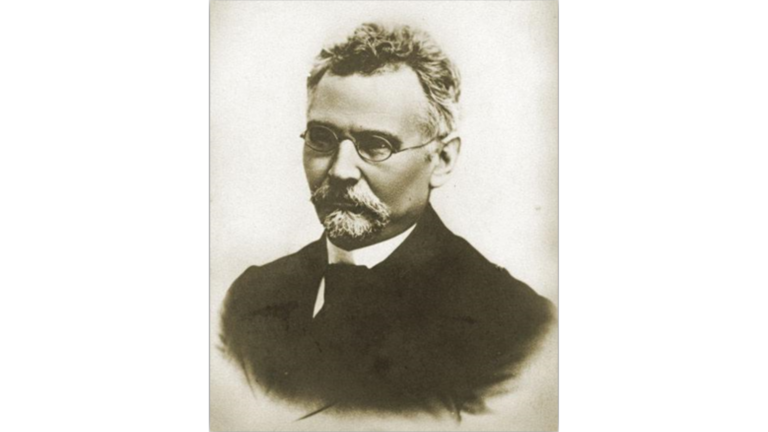 Portret czarno-biały (w odcieniach sepii) przedstawiający dojrzałego mężczyznę w ujęciu do pasa, zwróconego lekko w lewo, patrzącego prosto w obiektyw. Zdjęcie ma charakter formalny, studyjny, najprawdopodobniej wykonane w drugiej połowie XIX lub na początku XX wieku. Tło jest neutralne, jasne i gładkie – bez żadnych szczegółów, co kieruje całą uwagę na postać. Wygląd postaci: Wiek: Mężczyzna w wieku około 50–60 lat. Włosy: Krótkie, falowane, lekko uniesione, siwiejące po bokach, ciemniejsze na czubku głowy. Czoło: Szerokie, wysokie, częściowo zakryte kosmykiem włosów. Brwi: Gęste, ciemne. Oczy: Spokojne, poważne, patrzą prosto – ujęcie frontalne. Na oczach okrągłe okulary w cienkiej metalowej oprawce, typowe dla końca XIX wieku. Nos: Prosty, średniej wielkości. Wąsy i broda: Starannie przystrzyżone, siwe. Wąsy gęste, opadające po bokach ust. Pod brodą krótka, zaokrąglona bródka – łączy się z wąsami. Ubranie: Mężczyzna ubrany jest bardzo elegancko, w ciemną, dwurzędową marynarkę o klasycznym kroju. Pod szyją widoczny biały, wykrochmalony kołnierzyk koszuli oraz ciemny krawat lub muszka. Styl ubioru jest bardzo formalny, typowy dla uczonych, lekarzy lub urzędników z epoki. Wrażenie ogólne: Mężczyzna sprawia wrażenie osoby poważnej, wykształconej i dostojnej. Sposób w jaki jest ubrany i sfotografowany wskazuje na osobę o wysokiej pozycji społecznej lub naukowej, być może nauczyciela, naukowca lub dyrektora szkoły. Wyraz twarzy spokojny, skupiony, pozbawiony uśmiechu, ale nie surowy. Styl fotografii i charakter portretu przypominają zdjęcia zamieszczane w starych kronikach szkolnych, monografiach naukowych lub na tablicach pamiątkowych.