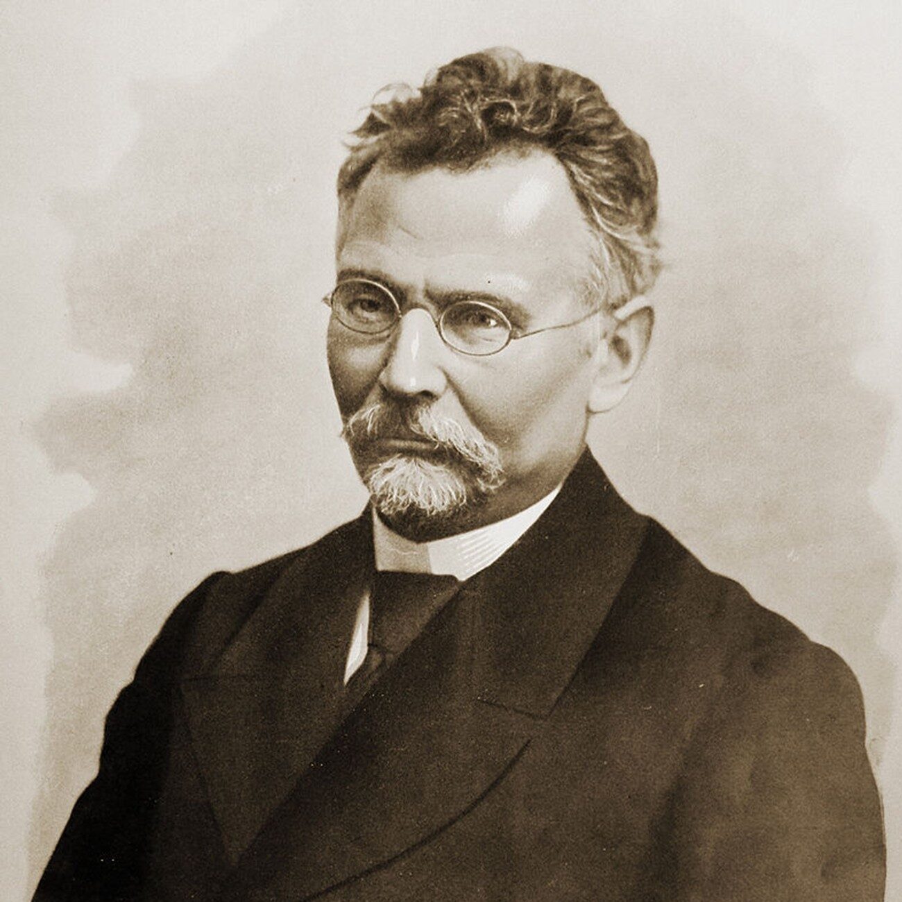 Portret czarno-biały (w odcieniach sepii) przedstawiający dojrzałego mężczyznę w ujęciu do pasa, zwróconego lekko w lewo, patrzącego prosto w obiektyw. Zdjęcie ma charakter formalny, studyjny, najprawdopodobniej wykonane w drugiej połowie XIX lub na początku XX wieku. Tło jest neutralne, jasne i gładkie – bez żadnych szczegółów, co kieruje całą uwagę na postać. Wygląd postaci: Wiek: Mężczyzna w wieku około 50–60 lat. Włosy: Krótkie, falowane, lekko uniesione, siwiejące po bokach, ciemniejsze na czubku głowy. Czoło: Szerokie, wysokie, częściowo zakryte kosmykiem włosów. Brwi: Gęste, ciemne. Oczy: Spokojne, poważne, patrzą prosto – ujęcie frontalne. Na oczach okrągłe okulary w cienkiej metalowej oprawce, typowe dla końca XIX wieku. Nos: Prosty, średniej wielkości. Wąsy i broda: Starannie przystrzyżone, siwe. Wąsy gęste, opadające po bokach ust. Pod brodą krótka, zaokrąglona bródka – łączy się z wąsami. Ubranie: Mężczyzna ubrany jest bardzo elegancko, w ciemną, dwurzędową marynarkę o klasycznym kroju. Pod szyją widoczny biały, wykrochmalony kołnierzyk koszuli oraz ciemny krawat lub muszka. Styl ubioru jest bardzo formalny, typowy dla uczonych, lekarzy lub urzędników z epoki. Wrażenie ogólne: Mężczyzna sprawia wrażenie osoby poważnej, wykształconej i dostojnej. Sposób w jaki jest ubrany i sfotografowany wskazuje na osobę o wysokiej pozycji społecznej lub naukowej, być może nauczyciela, naukowca lub dyrektora szkoły. Wyraz twarzy spokojny, skupiony, pozbawiony uśmiechu, ale nie surowy. Styl fotografii i charakter portretu przypominają zdjęcia zamieszczane w starych kronikach szkolnych, monografiach naukowych lub na tablicach pamiątkowych.
