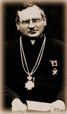 Na fotografii przedstawiono duchownego katolickiego, ujętego od pasa w górę. Zdjęcie jest czarno-białe, w stylu sepii, co nadaje mu historycznego, archiwalnego charakteru. Mężczyzna jest w średnim wieku lub starszy. Ma okrągłą twarz, łagodne rysy i lekki uśmiech na ustach. Nos ma średniej wielkości, oczy niewielkie, spoglądające lekko w bok (na lewo z perspektywy widza), okolone okularami w cienkiej, metalowej oprawce. Ma krótkie, ciemne włosy zaczesane do góry i na bok, z wyraźnie zaznaczoną linią przedziałka po prawej stronie głowy. Ubrany jest w ciemne, duchowne szaty – sutannę lub habit – bez widocznych ozdób na materiale. Na piersi nosi dwa długie, ozdobne łańcuchy, zawieszone symetrycznie na szyi i opadające po obu stronach. Na ich końcach widoczne są duże, bogato zdobione odznaczenia lub insygnia – jeden z nich ma kształt dużej, promienistej gwiazdy lub krzyża (może to być order), a drugi to mniejszy medal przypięty po lewej stronie klatki piersiowej. Odznaczenia wskazują na wysoką pozycję duchownego, być może zasługi społeczne, kościelne lub państwowe. Duchowny trzyma w rękach niezidentyfikowany biały przedmiot – może to być kartka papieru, dokument lub książeczka. Jego dłonie są splecione przed sobą, widoczne tylko częściowo. Tło zdjęcia jest jednolite i ciemne, co sprawia, że postać jest dobrze wyeksponowana i stanowi centralny punkt kompozycji. Krawędzie fotografii są delikatnie rozmyte, co podkreśla jej wiek i archiwalny charakter. Ogólne wrażenie: fotografia przedstawia spokojnego, godnego zaufania duchownego, którego postawa i odznaczenia sugerują zasłużone miejsce w historii Kościoła lub społeczeństwa. Styl zdjęcia i strój wskazują, że pochodzi ono z pierwszej połowy XX wieku.