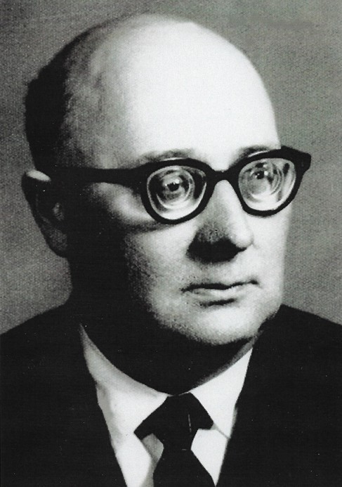 Czarno-białe zdjęcie portretowe, wykonane w stylu klasycznym, przedstawiające mężczyznę w średnim lub starszym wieku. Ujęcie sięga nieco poniżej ramion i ukazuje postać w półprofilu, zwróconą lekko w prawo (z punktu widzenia widza), przy jednoczesnym utrzymaniu wzroku skierowanego przed siebie. Opis postaci: Mężczyzna ma okrągłą, pełną twarz. Włosy bardzo rzadkie, niemal całkowicie łysy na czubku głowy, z krótkimi włosami po bokach i z tyłu. Brwi stosunkowo cienkie i jasne. Oczy duże, szeroko otwarte, co nadaje spojrzeniu nieco zaskoczony lub uważny wyraz. Ma wyraźne okulary o grubych, ciemnych oprawkach – okrągłe lub lekko owalne, bardzo charakterystyczne, wręcz dominujące w odbiorze twarzy. Nos dość duży, prosty, o zaokrąglonym czubku. Usta średniej wielkości, zamknięte, z neutralnym wyrazem. Broda okrągła, bez zarostu. Skóra jasna, gładka – bez widocznych zmarszczek, choć może to wynikać z jakości zdjęcia. Ubrany w formalny ciemny garnitur, białą koszulę i ciemny krawat z klasycznym węzłem. Tło i oświetlenie: Tło jednolite, w jasnoszarym odcieniu – nie odciąga uwagi od postaci. Oświetlenie miękkie, równomierne, dobrze doświetlające całą twarz, bez głębokich cieni. Charakter zdjęcia: Zdjęcie formalne, najprawdopodobniej portret urzędowy, naukowy lub dokumentacyjny. Styl opraw okularów może sugerować okres od lat 50. do 70. XX wieku. Mężczyzna wygląda poważnie