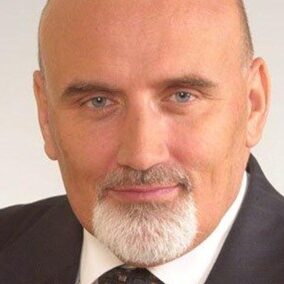 Zdjęcie portretowe, kolorowe, przedstawiające mężczyznę w średnim wieku lub nieco starszego – ujęcie typu "headshot", czyli pokazujące wyłącznie głowę i górną część ramion. Mężczyzna patrzy prosto w obiektyw, z lekkim, pewnym siebie uśmiechem. Tło jest jasne, jednolite, neutralne – prawdopodobnie białe lub bardzo jasno szare – co dodatkowo eksponuje twarz. Opis wyglądu osoby: • Głowa i twarz: o Mężczyzna ma łysinę, brak włosów na czubku głowy i bokach. o Zarost jest bardzo charakterystyczny: ma gęstą, krótką, siwą brodę, która pokrywa podbródek i część policzków, oraz siwe wąsy, które łączą się z brodą. Górna część brody jest delikatnie przyciemniona – może sugerować resztki pigmentu. o Twarz jest owalna, z wyraźnie zaznaczonymi kośćmi policzkowymi i symetrycznymi rysami. Skóra jest jasna, cera gładka, bez widocznych skaz. • Oczy i mimika: o Oczy są niebieskie, średniej wielkości, wyraziste, o łagodnym, ale zdecydowanym spojrzeniu. o Brwi są średniej grubości, dobrze uformowane, lekko podkreślające mimikę. o Kąciki ust delikatnie uniesione, co daje wrażenie spokojnego, pewnego siebie i profesjonalnego wyrazu twarzy. Ubiór: • Mężczyzna ubrany jest bardzo elegancko – ma na sobie ciemny garnitur, białą koszulę oraz krawat w odcieniach brązu lub szarości z delikatnym wzorem. • Krawat jest poprawnie zawiązany, garnitur dobrze dopasowany – co sugeruje dbałość o profesjonalny wizerunek. Atmosfera zdjęcia: • Zdjęcie ma charakter oficjalny lub biznesowy – mogłoby zostać użyte na stronie internetowej firmy, w materiałach promocyjnych, profilach zawodowych (np. LinkedIn) lub w publikacjach naukowych. • Osoba na zdjęciu prezentuje się jako doświadczony specjalista, lider lub menedżer, wzbudzający zaufanie i autorytet.