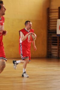 Na zdjęciu widać mężczyznę w średnim wieku, aktywnie grającego w koszykówkę na hali sportowej. Ubrany jest w czerwony, sportowy strój koszykarski – bezrękawnik i krótkie spodenki. Na jego stroju widnieje logo „VIP Agencja Reklamy” oraz numer „13” w kolorze białym, zarówno na koszulce, jak i na spodenkach. Strój ma biało-czarne lampasy po bokach. Mężczyzna nosi białe sportowe skarpetki oraz białe buty koszykarskie z ciemnymi detalami. Ma krótkie, ciemne włosy, lekki zarost (bródka), jest szczupły i w ruchu – biegnie lekko pochylony do przodu, kozłując piłkę koszykową (biało-czerwoną z szarymi przetarciami) prawą ręką.

Tło zdjęcia to sala gimnastyczna o drewnianej podłodze i żółtawych ścianach. Po prawej stronie widać drewniane drabinki gimnastyczne, częściowo zasłonięte banerem. W lewym górnym rogu kadru widoczna jest sylwetka drugiego zawodnika (rozmyta) w czerwonym stroju, ale twarz nie jest w pełni widoczna – tylko jego ramię i tors. Cała scena ma charakter dynamiczny – to fragment meczu lub treningu koszykówki halowej.
