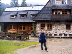 Zdjęcie przedstawia mężczyznę stojącego na tle dużego, górskiego schroniska zbudowanego z drewna i kamienia. Budynek ma rustykalny charakter – dolna część wykonana jest z nieregularnych kamieni w odcieniach beżu, szarości i różu, z drewnianymi oknami. Górna część to grube, drewniane bale z ciemnym, niemal czarnym gontowym dachem. Na poddaszu widać trzy małe okna z daszkami oraz jedno większe z balkonem. Na dachu zainstalowano panele słoneczne.

Mężczyzna stoi na środku placu, przed wejściem – jest starszy, ma krótkie siwe włosy i wąsy. Ubrany jest w granatową kurtkę, ciemne spodnie i sportowe buty. Ma na plecach mały plecak, dłonie schowane w kieszeniach. Wyraz jego twarzy jest pogodny i spokojny.

Plac przed schroniskiem jest nieco błotnisty, z kałużami i kamieniami, z lewej strony widać drewniane stoły piknikowe i ławki. W tle znajdują się gęste drzewa iglaste, otulone lekką mgłą – typowy krajobraz górski.