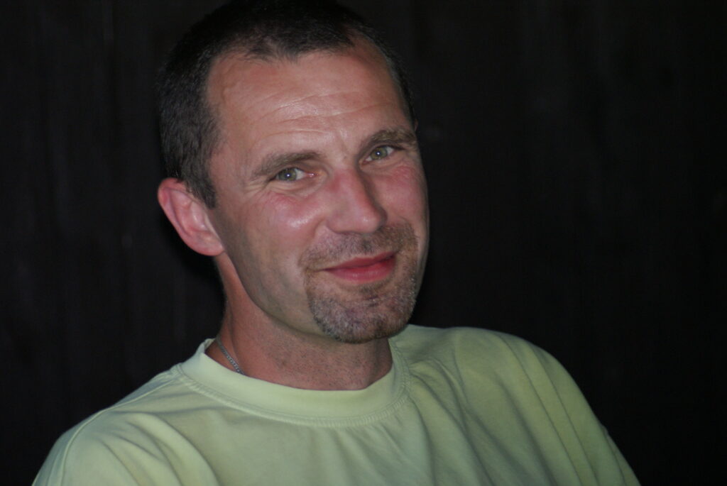 Na zdjęciu widoczny jest mężczyzna sfotografowany w średnim zbliżeniu, w kadrze od klatki piersiowej w górę. Mężczyzna ma około 35–45 lat, krótko ostrzyżone ciemne włosy i zarost w formie krótkiej bródki z wąsem, lekko przyprószonej siwizną. Ma jasną cerę i wyraźnie zarysowane rysy twarzy – wysokie czoło, ostro zarysowane kości policzkowe, delikatne zmarszczki mimiczne przy oczach. Patrzy bezpośrednio w obiektyw aparatu z ciepłym, łagodnym uśmiechem – jego usta są lekko uniesione, a oczy rozświetlone, co nadaje mu przyjazny i pogodny wyraz twarzy. Ubrany jest w jasną, pastelowo-żółtą koszulkę z krótkim rękawem, z okrągłym dekoltem. Na szyi ma cienki srebrny łańcuszek, który częściowo wystaje spod koszulki. Tło zdjęcia jest jednolite, bardzo ciemne – niemal czarne – i nieostre, co powoduje, że postać jest mocno wyeksponowana i dobrze oświetlona na pierwszym planie. Oświetlenie wydaje się być sztuczne lub błysk flesza – twarz mężczyzny jest równomiernie oświetlona, a cienie są minimalne. Zdjęcie sprawia wrażenie wykonania w pomieszczeniu lub wieczorem. Atmosfera ujęcia jest kameralna, spokojna i pozytywna.
