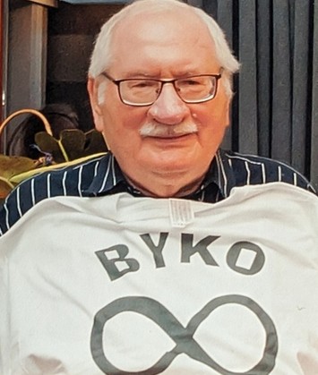 Na zdjęciu znajduje się starszy mężczyzna siedzący w pomieszczeniu. Jest uśmiechnięty i patrzy wprost na obiektyw aparatu. Ma siwe, rzadkie włosy i nosi okulary w prostokątnej oprawie. Jego twarz ma wyraźnie zarysowane rysy – nos jest prosty, policzki lekko zarumienione, a wąsy białe i starannie przycięte. Mężczyzna ma na sobie ciemną koszulę w pionowe białe paski, a na niej trzyma rozwieszony biały T-shirt. Koszulka zasłania jego tors i większość koszuli. Na białej koszulce nadrukowane są dwa elementy w kolorze czarnym lub ciemnoszarym: W górnej części nadrukowane jest słowo "BYKO" – napisany dużymi, pogrubionymi literami bezszeryfowymi (czcionka bez ozdobników). Poniżej, centralnie, znajduje się duży symbol nieskończoności – czyli pozioma ósemka (∞), również w ciemnym kolorze. Tło zdjęcia jest nieco przyciemnione i zamazane – widoczna jest tapicerowana, ciemna ściana lub zasłona po prawej stronie oraz fragment jasnego przedmiotu przypominającego torbę lub torebkę po lewej stronie. Cała scena jest dobrze oświetlona, co pozwala dostrzec szczegóły twarzy i ubioru mężczyzny. Symbolika: – Słowo „BYKO” oraz znak nieskończoności mogą mieć symboliczne znaczenie – być może to nazwa drużyny, grupy, pseudonim lub odniesienie do siły i trwałości (byk jako symbol siły, nieskończoność jako nieprzemijalność).