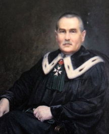 Na obrazie znajduje się starszy mężczyzna w dostojnym stroju ceremonialnym, siedzący na ciemnym krześle z podłokietnikami. Kompozycja ma charakter formalnego portretu olejnego – postać jest ukazana w pozycji siedzącej, frontalnie, z lekkim skrętem w prawo (z perspektywy widza). Tło jest ciemne, neutralne, lekko fakturalne, bez szczegółów – skupia uwagę całkowicie na przedstawionej osobie. Wygląd postaci: Mężczyzna ma prostą postawę, patrzy spokojnie przed siebie. Twarz jest szczupła, z wysokim czołem, prostym nosem, cienkimi wargami i wyraźnie zarysowanymi kośćmi policzkowymi. Cera jasna, lekko zaróżowiona, skóra dojrzała, ale gładka jak na wiek postaci – prawdopodobnie około 60–70 lat. Włosy są krótko ostrzyżone, szpakowate, czyli ciemnoszare z przewagą siwizny, szczególnie na skroniach. Wąsy starannie przystrzyżone, również siwe, równe i niezbyt gęste. Brwi ciemniejsze, lekko uniesione. Strój: Mężczyzna nosi czarną togę – długi, luźny strój o szerokich rękawach, charakterystyczny dla profesorów uniwersyteckich lub dostojników kościelnych. Na szyi widnieje ceremonialna, ozdobna kolara (kołnierz), w kształcie litery „V”, wyłożona białym futrem z czarnymi akcentami – typowy element stroju akademickiego lub sądowego. Na piersi widoczna jest wstęga z zielonego materiału, do której przypięty jest duży order w formie białego krzyża z czarną obwódką, przypominający krzyż maltański, na tle czarnej gwiazdy. Nad nim znajduje się ozdobny, czerwony emblemat – być może rozeta lub herb. Całość przypięta do zielonej wstążki złożonej w fałdy. Dłonie widoczne są częściowo – jedna spoczywa luźno na kolanie, druga jest schowana w rękawie lub na podłokietniku. Mężczyzna siedzi prosto, z godnością i spokojem, z lekko zaciśniętymi ustami. Charakterystyka ogólna: Styl obrazu wskazuje na realistyczne malarstwo portretowe, wykonane z dbałością o szczegóły ubioru i godność postaci. Przedstawiona osoba jest prawdopodobnie profesorem, rektorem uczelni lub wysokim rangą urzędnikiem państwowym lub prawnikiem – z uwagi na strój ceremonialny i odznaczenia. Obraz oddaje atmosferę szacunku, powagi i dostojeństwa.