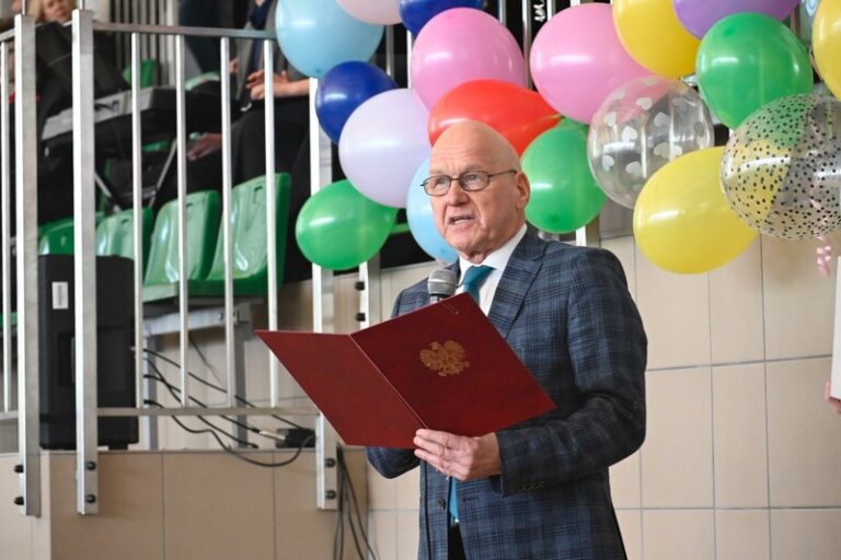 Na zdjęciu widoczny jest mężczyzna w średnim wieku, który przemawia do mikrofonu w trakcie uroczystości wewnątrz budynku – prawdopodobnie w hali sportowej lub sali gimnastycznej przystosowanej na wydarzenie szkolne. Mężczyzna jest łysy, nosi okulary i ma na sobie elegancki garnitur w ciemnoniebieską kratę oraz białą koszulę i niebieski krawat. Trzyma w dłoniach bordową teczkę lub ozdobny folder z godłem Polski (orzeł w koronie) na przedniej okładce – wygląda na to, że czyta z niego przemówienie. Tło zdjęcia zdobi wiele kolorowych balonów – są w odcieniach żółtego, zielonego, czerwonego, różowego, fioletowego, niebieskiego, przezroczyste z białymi kropkami oraz złote w czarne kropki. Balony są gęsto zawieszone i tworzą kolorowy, radosny klimat. Za mężczyzną widać część widowni – są tam zielone, plastikowe siedzenia trybun oraz barierki z metalowych rur. Część osób siedzi na tych miejscach, jednak są widoczne tylko ich fragmenty – dłonie, kolana lub fragmenty sylwetek. Ściany pomieszczenia wyłożone są jasnymi kafelkami w kolorze beżowym, co sugeruje, że może to być pomieszczenie używane na co dzień w celach sportowych, ale dostosowane do uroczystości. Uroczystość może być zakończeniem roku szkolnego, świętem szkoły, pasowaniem lub innym oficjalnym wydarzeniem edukacyjnym.