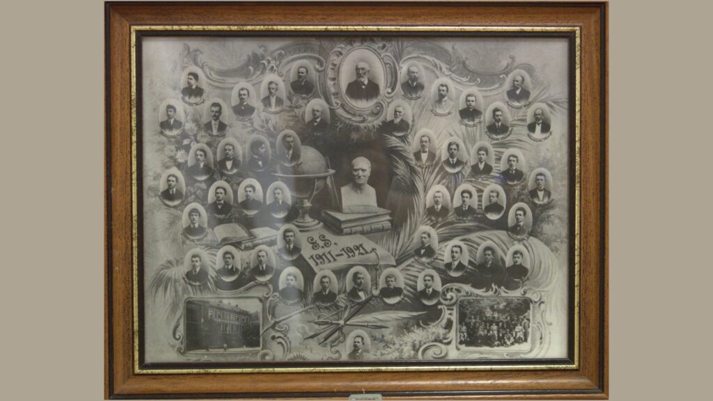 Zdjęcie przedstawia ozdobną, czarno-białą tablicę pamiątkową z portretami popiersiowymi uczniów i nauczycieli z lat 1911–1921. Kompozycja ma charakter secesyjny – portrety umieszczone są w ozdobnych ramkach owalnych i prostokątnych. W centrum znajduje się popiersie mężczyzny ustawione na książkach, obok globus i inne symbole edukacji. W tle – motywy liści palmowych i ozdobników roślinnych. Na dole duży napis „S.S. 1911–1921”. W lewym dolnym rogu zdjęcie budynku szkoły, w prawym dolnym – grupowe zdjęcie uczniów. Całość zamknięta w grubej, drewnianej ramie.