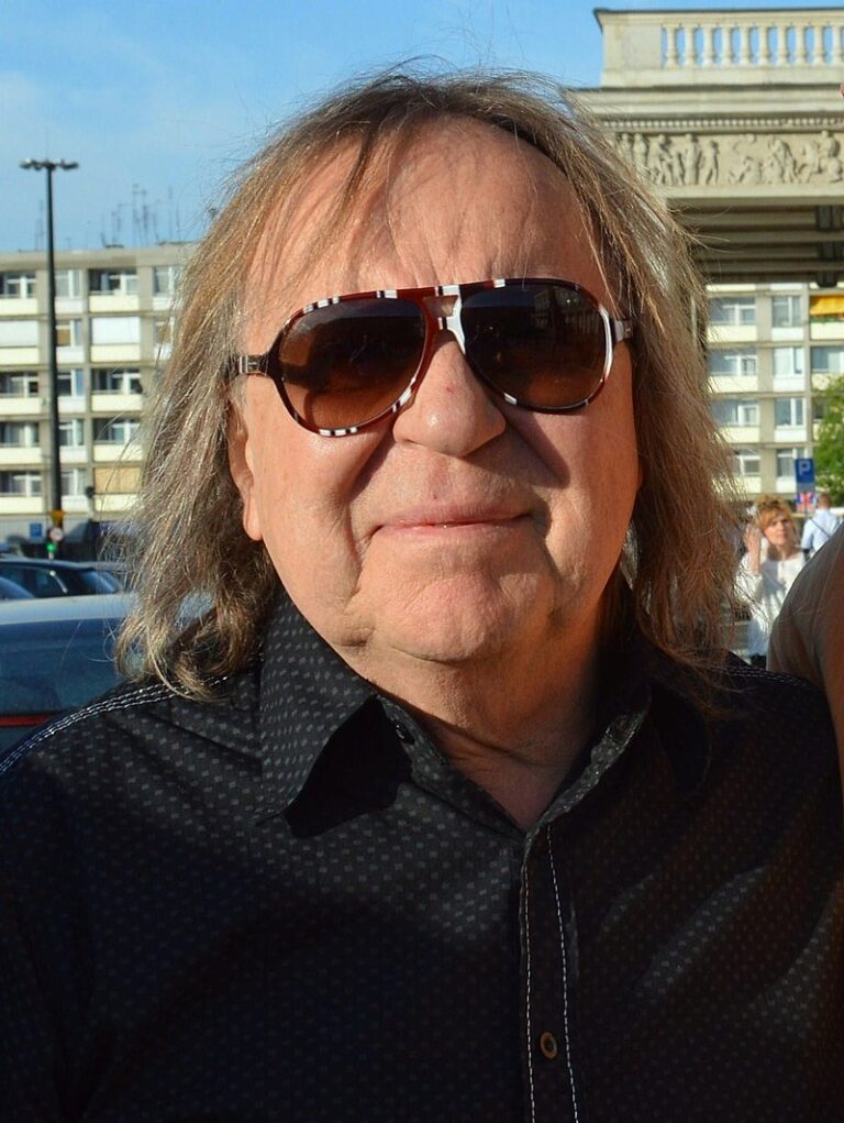 Na zdjęciu znajduje się starszy mężczyzna Romuald Lipko z długimi, jasnobrązowymi włosami sięgającymi ramion. Ma na sobie ciemną koszulę w drobny wzór i nosi duże okulary przeciwsłoneczne w grubej oprawce z białym wzorem. Twarz mężczyzny jest pogodna, lekko uśmiechnięta. W tle widać fragment miasta: szary budynek mieszkalny z balkonami i budynek z kolumnami oraz płaskorzeźbą. Zdjęcie zostało wykonane w słoneczny dzień, najpewniej podczas wydarzenia plenerowego.