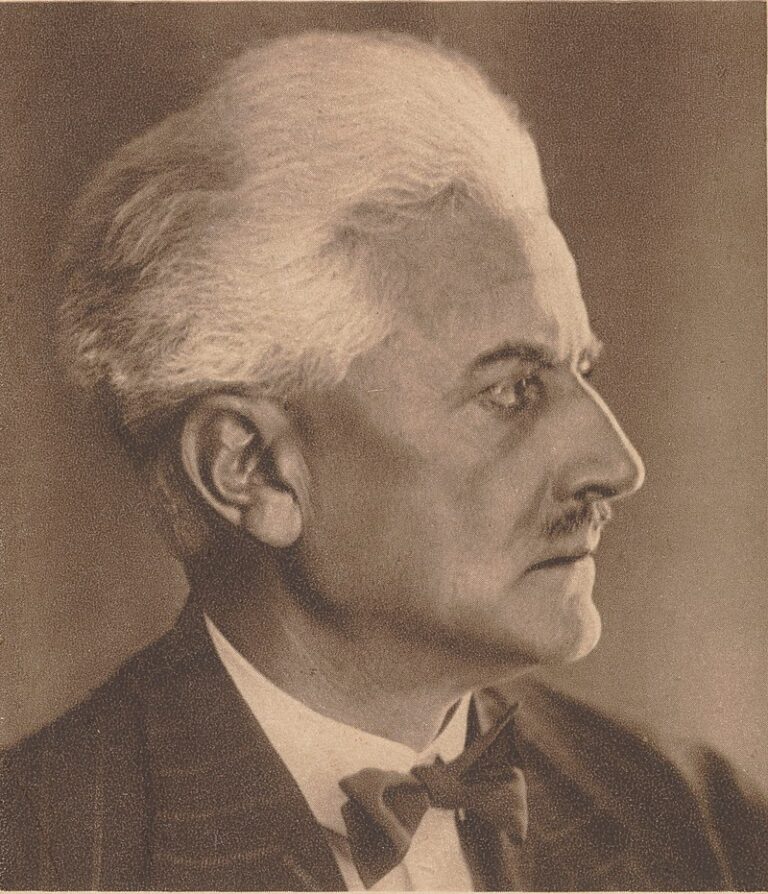 Czarno-biała fotografia portretowa Andrzeja Struga, pisarza i polityka, w klasycznej konwencji pocztówkowej. Mężczyzna ukazany z profilu – patrzy w prawo. Ma gęste, siwe włosy starannie uczesane do tyłu oraz starannie przycięty wąs. Ubrany jest w elegancki garnitur i muszkę. Tło neutralne, owalna ramka podkreśla oficjalny charakter zdjęcia. Styl i forma sugerują, że pochodzi z początku XX wieku.