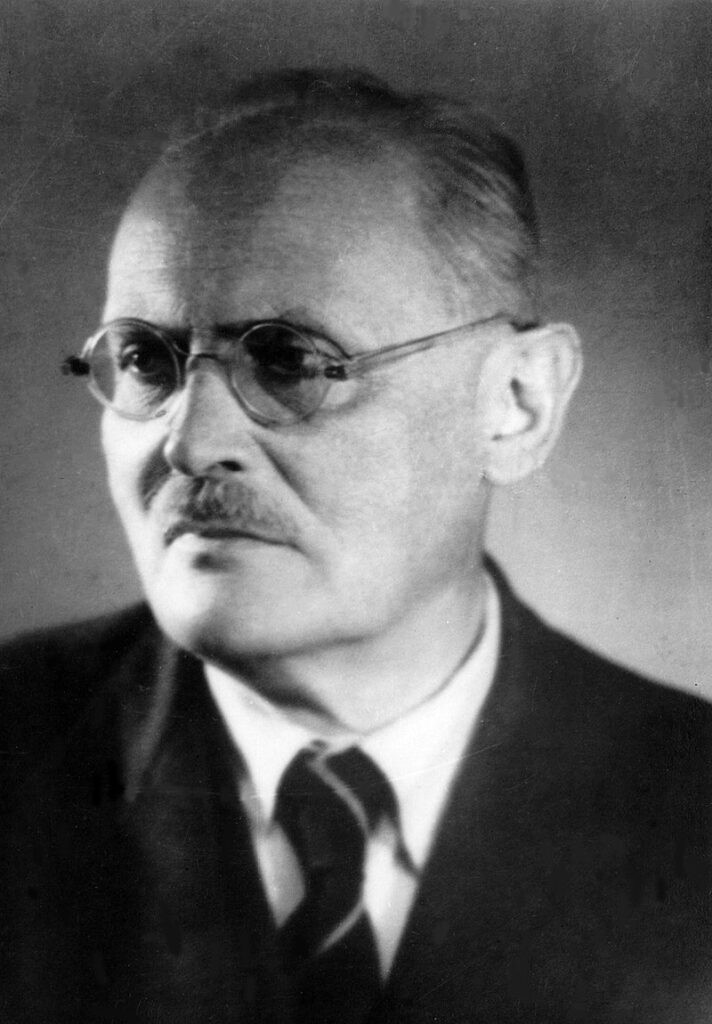 Brązowo-tonowany, profilowy portret Bolesława Hryniewieckiego, botanika i profesora. Starszy mężczyzna z wyrazistą białą fryzurą, zaczesaną do góry, oraz cienkim, zadbanym wąsem. Jego twarz jest poważna i skupiona. Ubrany w elegancki garnitur z muszką. Styl zdjęcia i jakość wskazują na lata 30. XX wieku. Tło rozmyte, ciepłe oświetlenie.