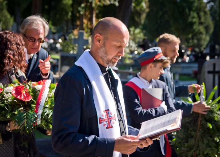 Na pierwszym planie widzimy księdza – łysego mężczyznę z krótkim zarostem, ubranego w ciemną kurtkę i białą stułę. W rękach trzyma otwartą książkę liturgiczną i czyta z niej modlitwę. W tle widać uczniów z pocztu sztandarowego oraz inne osoby, jedna z nich trzyma zielony wieniec.