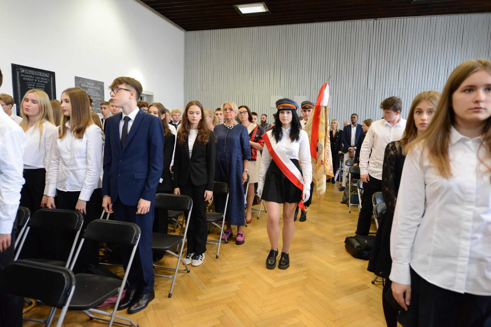 Na środku fotografii znajduje się uczennica w galowym stroju: biała koszula, czarna krótka spódnica, czapka szkolna i szarfa w barwach narodowych. Za nią dwie kolejne osoby niosą sztandar z orłem i napisem. Uczniowie po bokach w garniturach i białych koszulach stoją na baczność. Za nimi nauczyciele i rodzice. Sala jest pełna, w tle biało-szare ściany z pamiątkowymi tablicami.