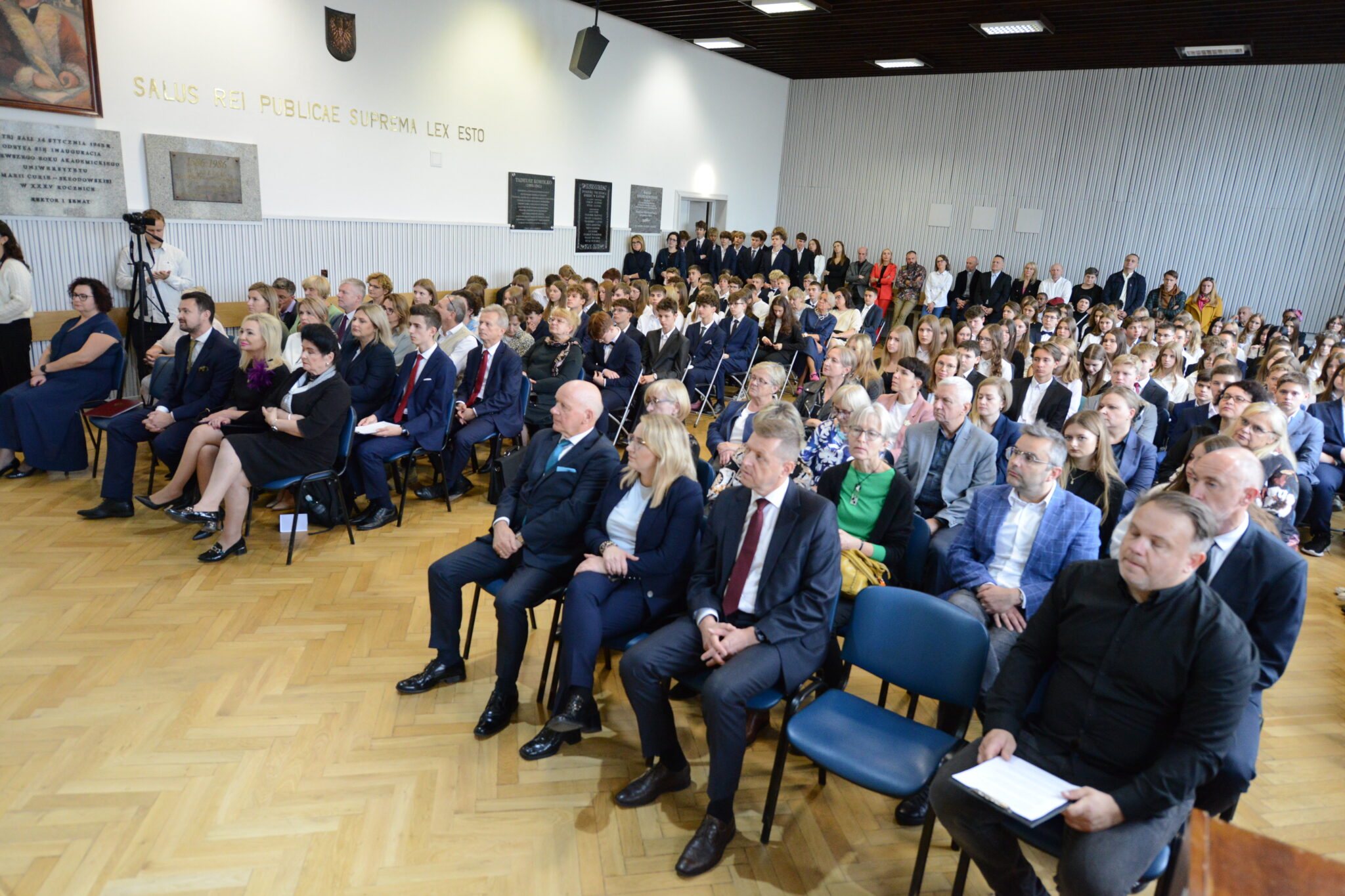 Setki osób siedzi w równo ustawionych rzędach. Uczniowie w eleganckich strojach — garnitury, białe koszule, spódnice, marynarki. Wśród nich nauczyciele, dyrekcja i zaproszeni goście. Z przodu sala wypełniona osobami dorosłymi, w dalszych rzędach młodzież szkolna. Na ścianie napis po łacinie „Salus rei publicae suprema lex esto”, a poniżej kilka tablic pamiątkowych.
