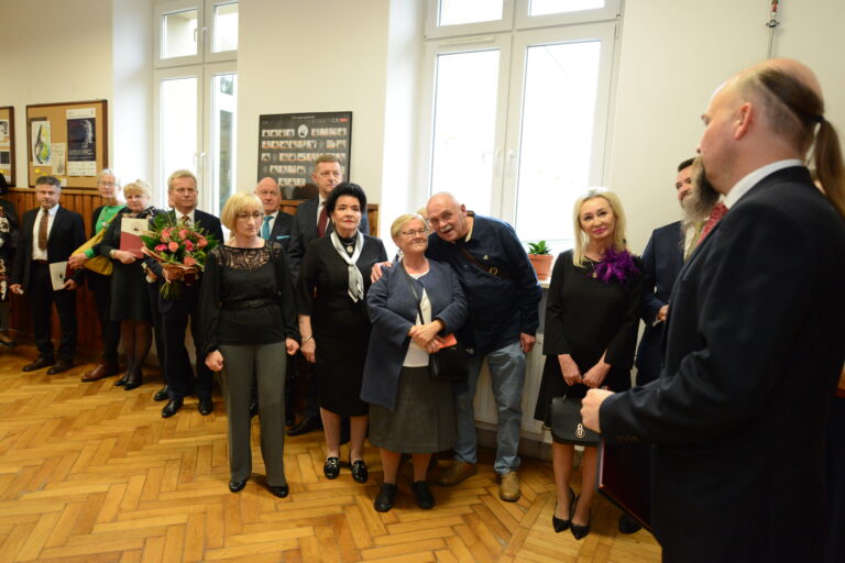 Zdjęcie przedstawia formalne spotkanie w przestrzeni szkolnej — prawdopodobnie korytarzu. Wnętrze jest jasne, z drewnianą podłogą w jodełkę, ścianami w kolorze kremowym i dwoma dużymi oknami w tle, przez które wpada dzienne światło. Na parapecie stoją dwie doniczki z zielonymi roślinami. W centrum zdjęcia zgromadziła się grupa około piętnastu osób, głównie starszych dorosłych, ubranych elegancko — w garnitury, sukienki, bluzki i marynarki. Wśród kobiet dominują czarne stroje wizytowe, niektóre z dodatkami w postaci broszek lub apaszek. Mężczyźni mają na sobie garnitury, niektórzy z krawatami. Jedna z kobiet trzyma bukiet różowo-zielonych kwiatów, co sugeruje, że mogła zostać uhonorowana. Niektóre osoby trzymają też teczki lub dokumenty. Po prawej stronie zdjęcia, częściowo odwrócony tyłem, stoi mężczyzna w garniturze z charakterystycznym uczesaniem — wygoloną głową po bokach i długim kucykiem z tyłu. Wydaje się przemawiać lub prowadzić uroczystość. Inne osoby patrzą na niego z uwagą i uprzejmym uśmiechem. Tło uzupełniają tablice informacyjne z plakatami oraz czarno-białe zdjęcia klasowe oprawione w ramki. Atmosfera spotkania jest ciepła, pełna szacunku i skupienia — najprawdopodobniej to spotkanie absolwentów lub uroczystość pożegnalna pracownika szkoły.