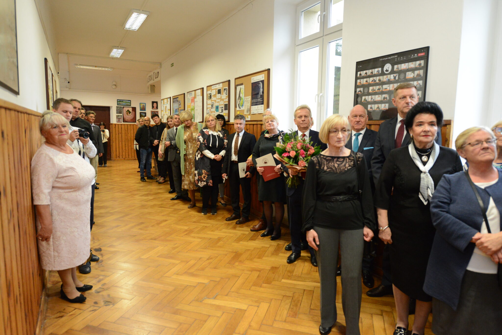 Grupa dorosłych i starszych osób, prawdopodobnie emerytowanych nauczycieli i absolwentów, zgromadzona na szkolnym korytarzu z drewnianą boazerią. W tle gazetki szkolne i tablice pamiątkowe. Kilka osób trzyma bukiety kwiatów, część trzyma teczki lub dokumenty. Na twarzach wyraz wzruszenia i wspomnienia. Jedna osoba z przodu trzyma ramieniem drugą w serdecznym geście.