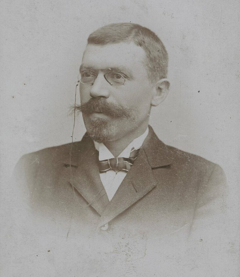 Stara fotografia portretowa Edmunda Biernackiego – mężczyzny z charakterystycznym dużym wąsem i krótkimi włosami. Nosi okulary typu pince-nez, przypięte sznurkiem do ucha. Ubrany jest w elegancki, ciemny garnitur, białą koszulę i muchę. Tło jasne i neutralne, styl zdjęcia formalny, wykonany zapewne w atelier pod koniec XIX wieku.