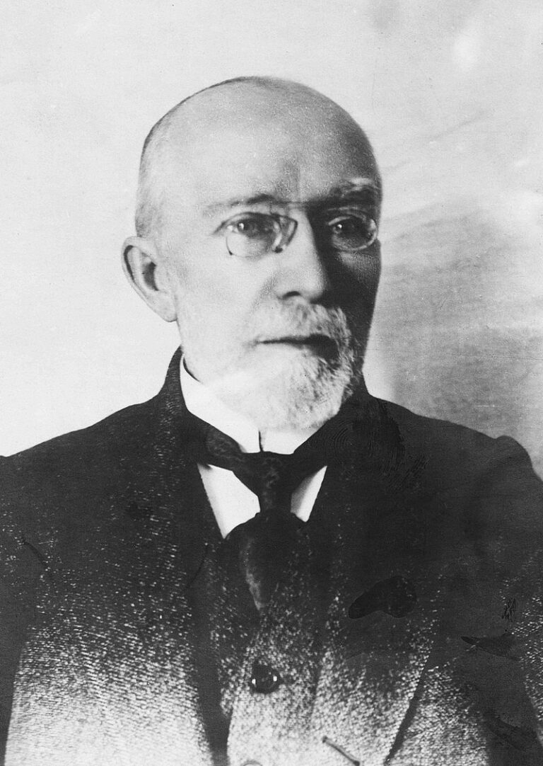 Czarno-biała fotografia Erazma Majewskiego – starszego mężczyzny z łysiną, siwą brodą i okularem pince-nez. Ubrany w garnitur z kamizelką i czarnym krawatem. Jego twarz ma łagodny, skupiony wyraz. Zdjęcie wykonano na jasnym tle, zapewne w pracowni fotograficznej. Styl sugeruje przełom XIX i XX wieku.