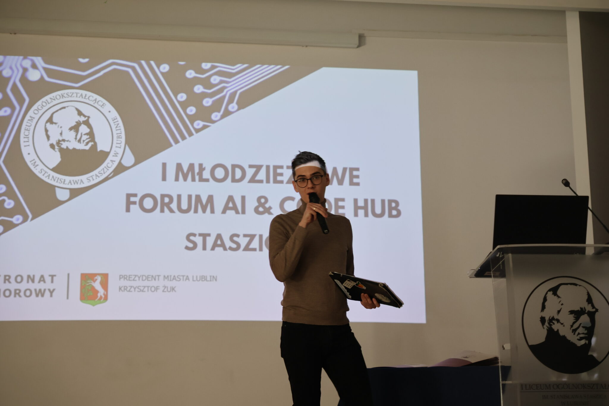 Mężczyzna z krótkimi ciemnymi włosami i okularami, ubrany w brązowy golf i czarne spodnie, trzyma mikrofon i przemawia do publiczności. W jednej ręce trzyma również laptopa z naklejkami (w tym jedna z grafiką brzoskwini). Tło stanowi biały ekran z dużym napisem w języku polskim: „I MŁODZIEŻOWE FORUM AI & CODE HUB – STASZIC 2025”. Na slajdzie znajduje się także logo szkoły oraz herb Miasta Lublin i napis: „Patronat Honorowy – Prezydent Miasta Lublin Krzysztof Żuk”.