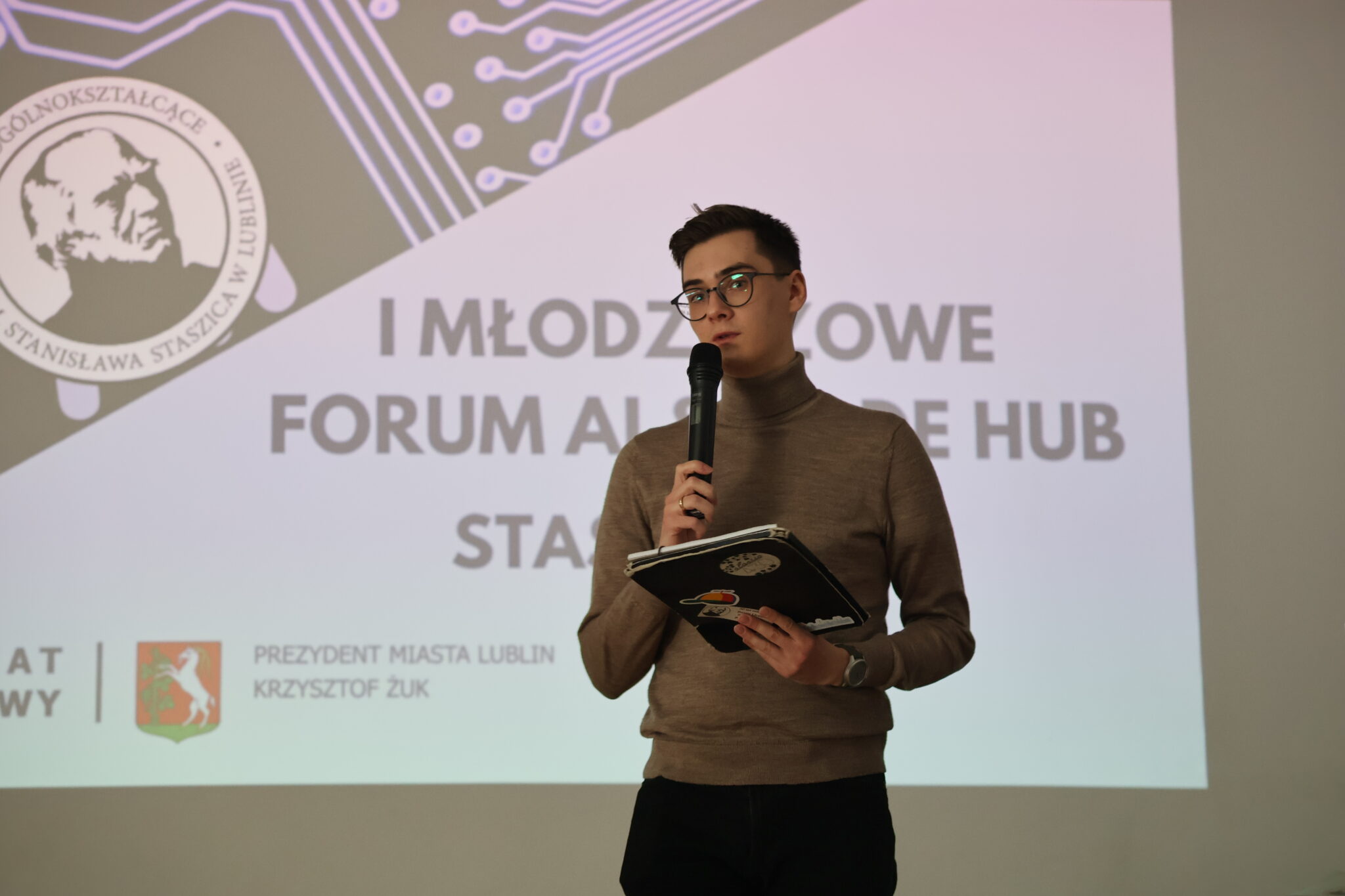 Mężczyzna z krótkimi ciemnymi włosami i okularami, ubrany w brązowy golf i czarne spodnie, trzyma mikrofon i przemawia do publiczności. W jednej ręce trzyma również laptopa z naklejkami (w tym jedna z grafiką brzoskwini). Tło stanowi biały ekran z dużym napisem w języku polskim: „I MŁODZIEŻOWE FORUM AI & CODE HUB – STASZIC 2025”. Na slajdzie znajduje się także logo szkoły oraz herb Miasta Lublin i napis: „Patronat Honorowy – Prezydent Miasta Lublin Krzysztof Żuk”.