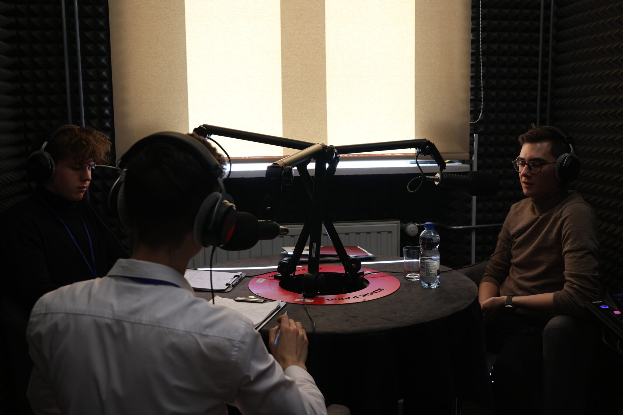 W pomieszczeniu wyciszonym panelami akustycznymi (ściany wyłożone czarną pianką dźwiękochłonną) znajduje się studio podcastowe. Przy okrągłym stole pokrytym czarnym obrusem siedzi trzech młodych mężczyzn w słuchawkach i przy mikrofonach. Jeden z nich (na zdjęciu 5 po prawej) nosi brązowy golf i okulary – ten sam, który występował wcześniej na scenie. Przed każdym z uczestników znajdują się notatki lub dokumenty. Na stole stoi również butelka wody i szklanka.