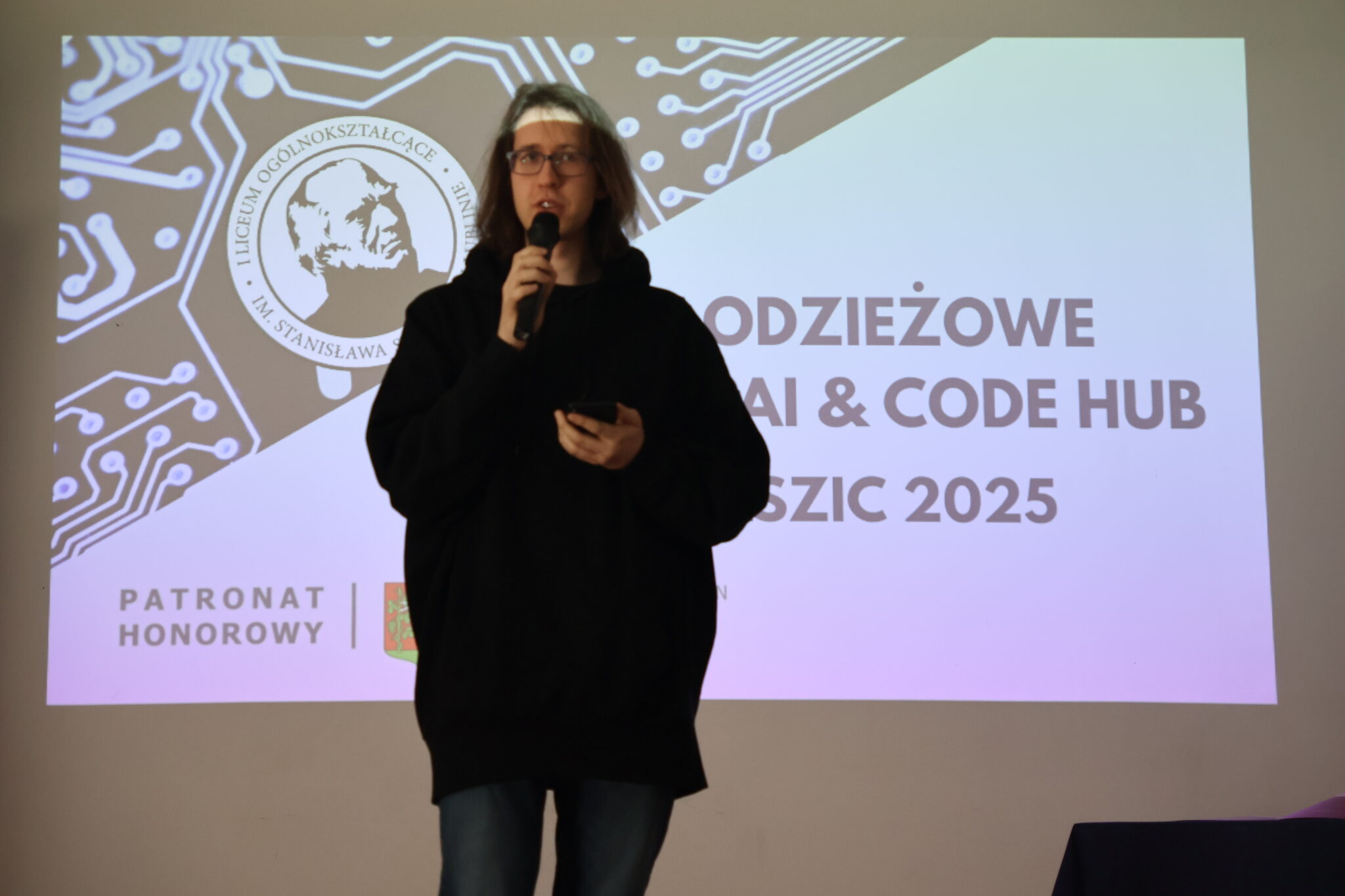 Chłopak w czarnej obszernej bluzie z kapturem, noszący okulary, trzyma mikrofon i telefon komórkowy. Stoi na tle tego samego slajdu co wcześniej – wydarzenia „Staszic 2025”. Wygląda na zamyślonego, być może odczytuje cytat lub przemówienie z telefonu.