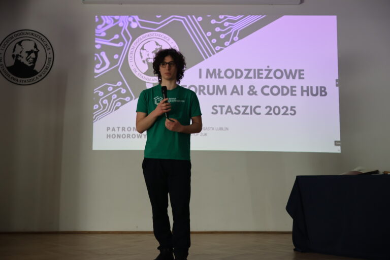 Zdjęcie przedstawia młodego chłopaka w wieku szkolnym, stojącego na scenie lub w sali prezentacyjnej. Chłopak ma jasną karnację, średniej długości ciemne, kręcone włosy sięgające do szyi. Nosi okulary z ciemną oprawką. Ubrany jest w zieloną koszulkę z krótkim rękawem z białym nadrukiem z przodu, na którym widnieje napis „AKADEMIA PROGRAMOWANIA” oraz geometryczny symbol przypominający połączenie sześcianów. Do tego ma ciemne spodnie i czarne buty. Trzyma mikrofon w obu dłoniach i patrzy prosto przed siebie, najpewniej przemawia lub prezentuje. Stoi na tle dużego ekranu projekcyjnego z wyświetlonym slajdem prezentacji. Na slajdzie widoczny jest tytuł wydarzenia: „I MŁODZIEŻOWE FORUM AI & CODE HUB STASZIC 2025” Pod spodem widnieje informacja o patronacie honorowym Prezydenta Miasta Lublina – Krzysztofa Żuka. Po lewej stronie slajdu znajduje się grafika przedstawiająca obwody drukowane oraz herb szkoły – „I LICEUM OGÓLNOKSZTAŁCĄCE IM. STANISŁAWA STASZICA W LUBLINIE”. Taki sam herb widoczny jest również fizycznie na ścianie po lewej stronie zdjęcia, w postaci okrągłej tablicy. Po prawej stronie zdjęcia, w dolnym rogu, znajduje się stolik lub podwyższenie nakryte ciemnym, granatowym obrusem – na nim leżą luźne papiery lub materiały konferencyjne. Całość utrzymana jest w formalnej, edukacyjnej atmosferze – wydarzenie wygląda na oficjalne forum technologiczne lub konkurs programistyczny dla młodzieży.