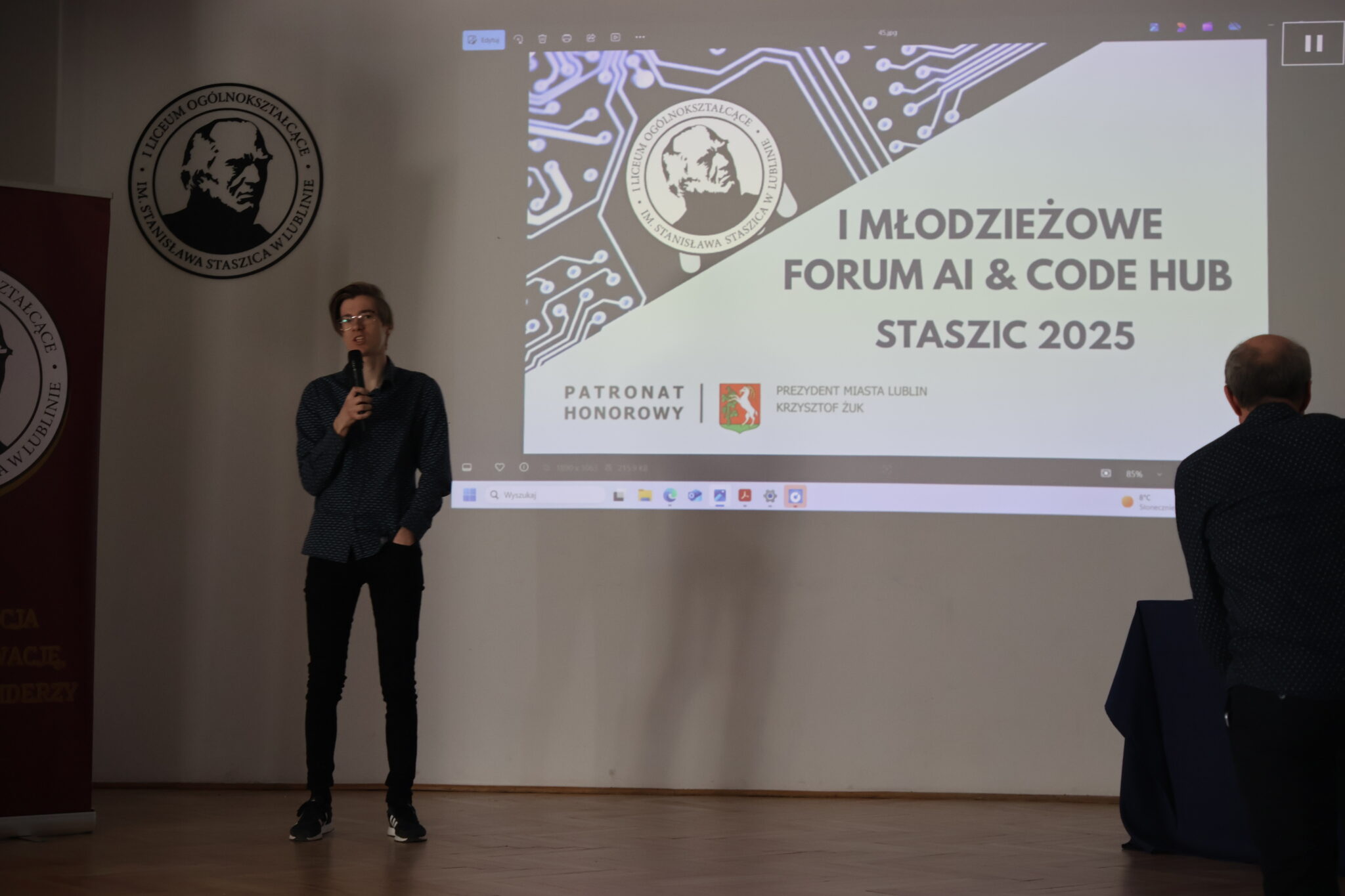 Na scenie stoi młody chłopak w okularach i ciemnej koszuli w drobny wzór. Ma rękę w kieszeni i mówi do mikrofonu, stojąc na tle ekranu z napisem „I MŁODZIEŻOWE FORUM AI & CODE HUB STASZIC 2025”. Po prawej stronie kadru widać sylwetkę starszego mężczyzny, który zdaje się przygotowywać do zejścia ze sceny lub przygląda się prezentacji.