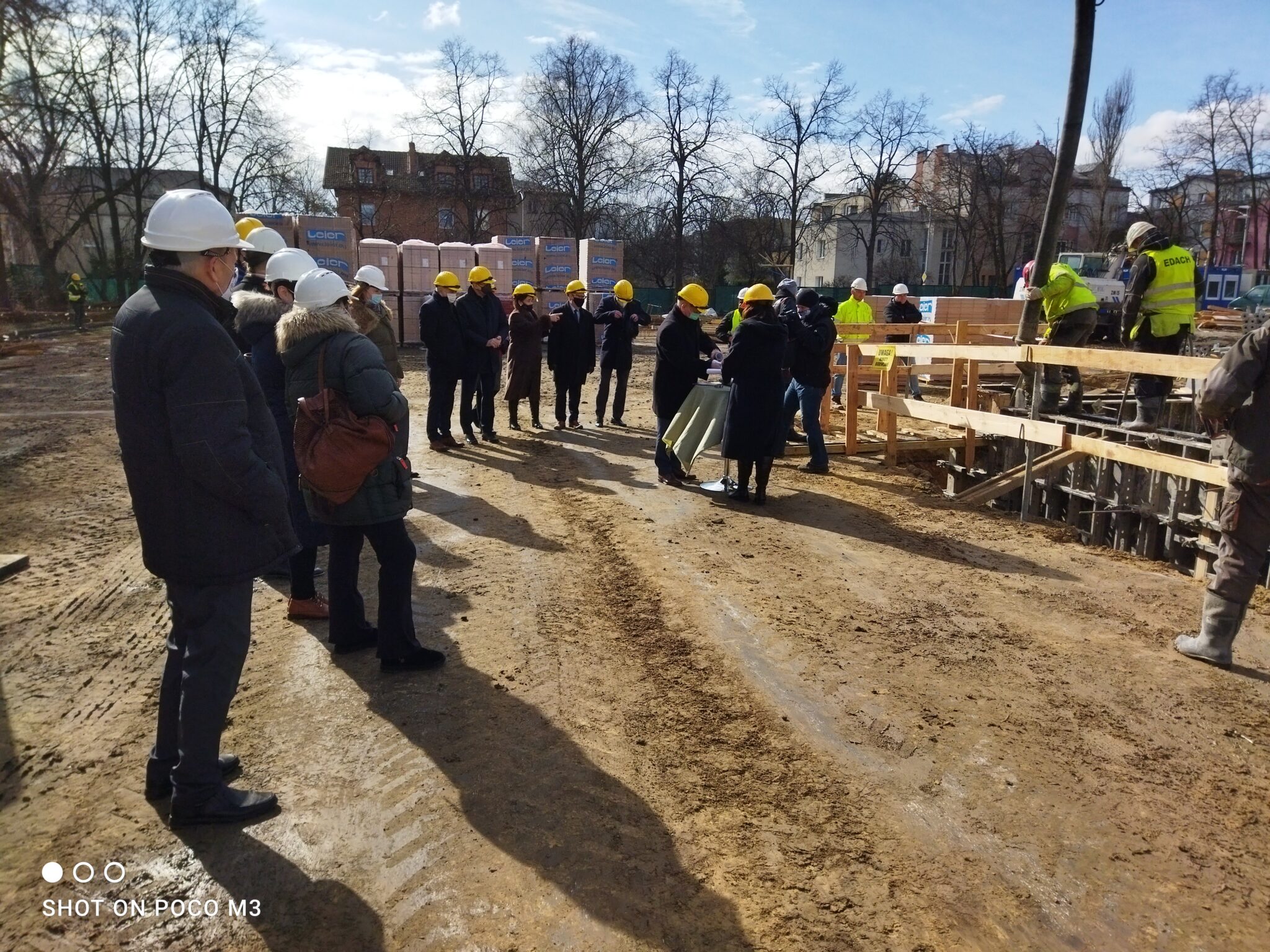 Ujęcie szerokie z perspektywy obserwatora stojącego po lewej stronie budowy. W centralnej części znajduje się grupa ludzi (około 15 osób) zebranych w półokręgu. Wszyscy noszą kaski ochronne (żółte i białe) oraz maseczki na twarzach. Po prawej stronie kilku robotników stoi na rusztowaniu nad wykopem, wlewając beton. W tle drzewa bez liści, budynki mieszkalne i palety materiałów budowlanych.