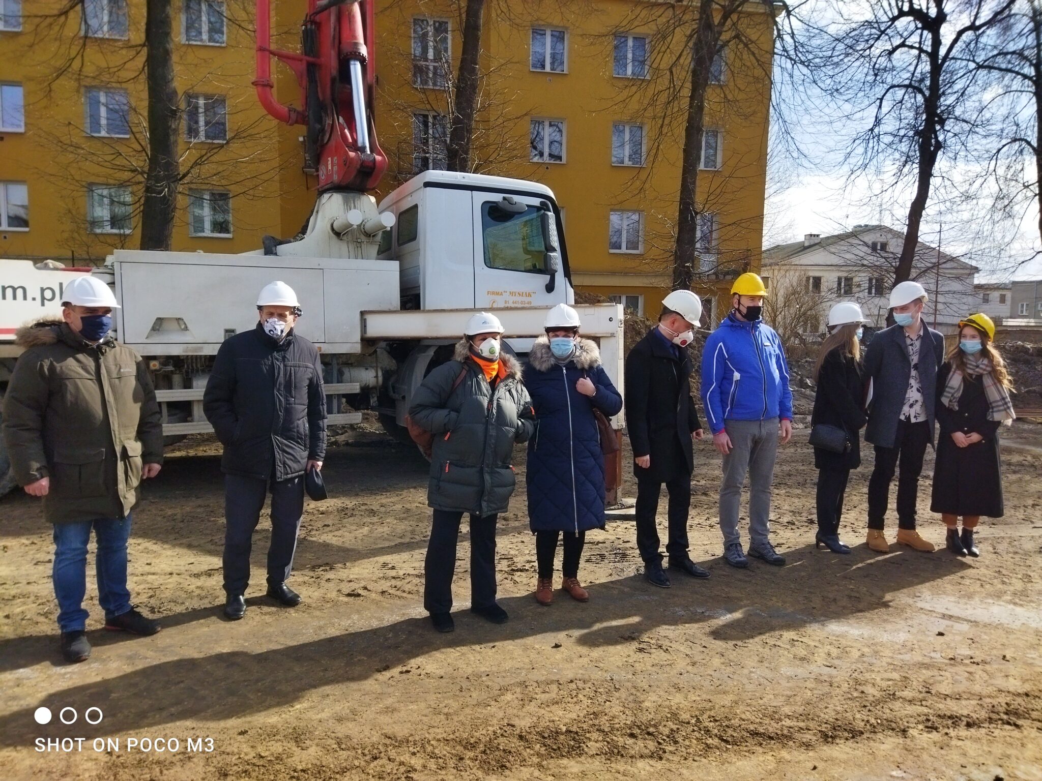 Grupa dziewięciu osób stoi przed dużą białą betoniarką z napisem „www.mysiak.com.pl”. Wszyscy noszą kaski i zimowe ubrania. Trzy osoby mają żółte kaski, pozostałe białe. Tło stanowi żółty budynek mieszkalny z wieloma oknami i balkonami, oraz drzewa bez liści.
