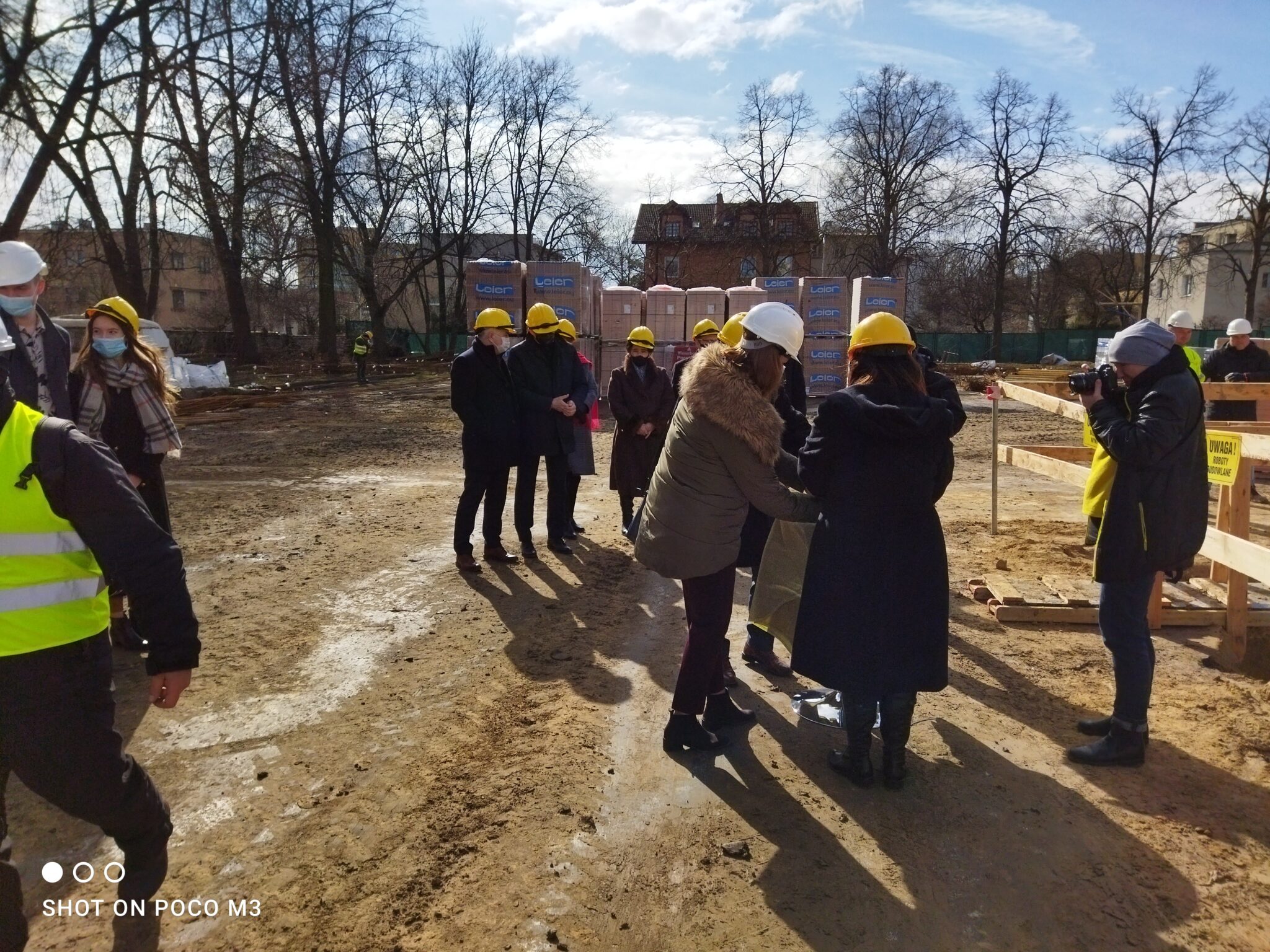 Zbliżenie grupy gości stojących wokół zielonego, okrągłego stolika z metalową tubą (symbolicznym kamieniem węgielnym). Dwie kobiety w ciemnych kurtkach i żółtych kaskach trzymają folię ochronną. Obok nich mężczyzna z aparatem fotograficznym robi zdjęcie wydarzeniu. W tle – palety materiałów firmy „Leier” i osoby w kaskach.