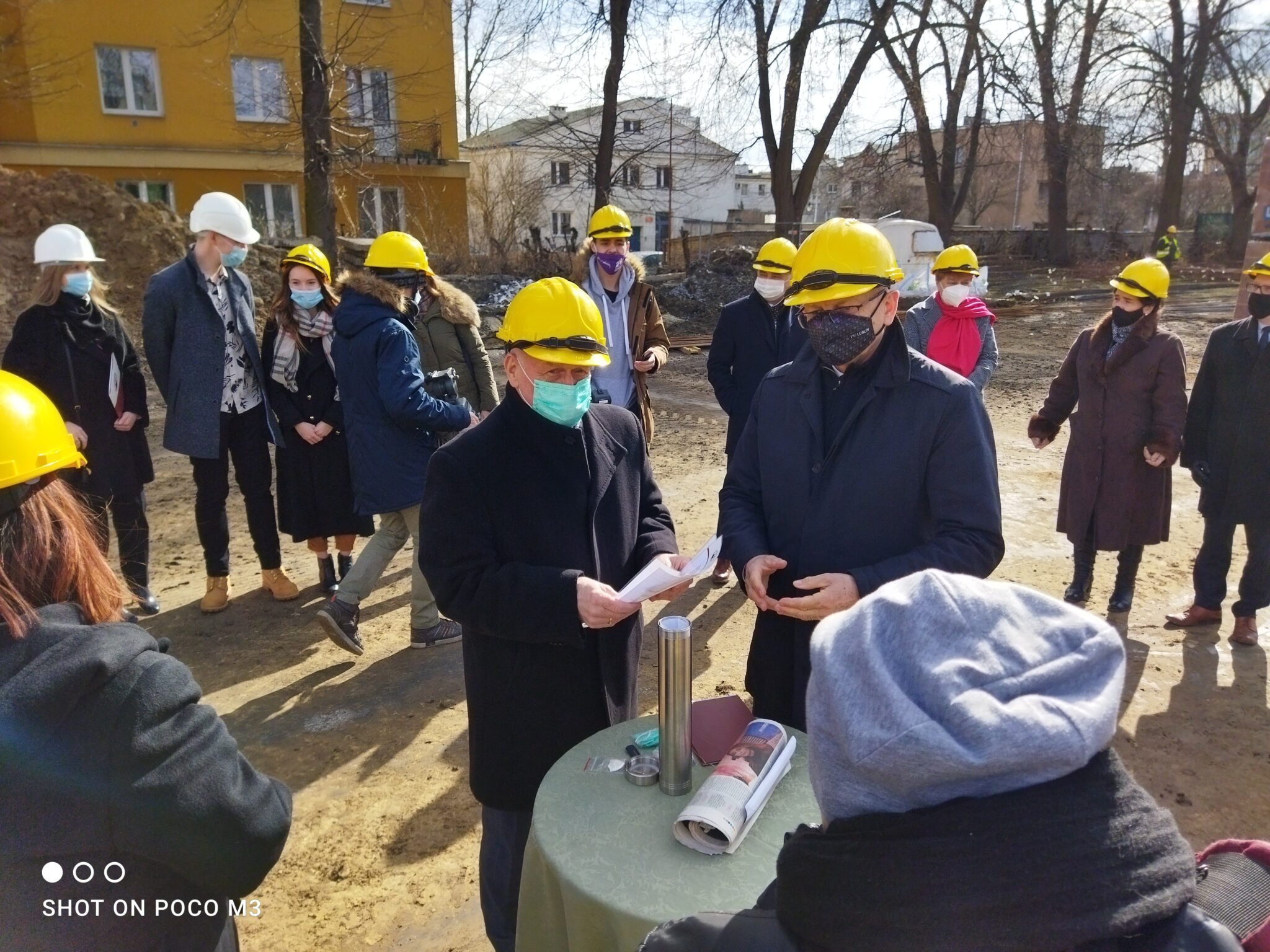 Kontynuacja poprzedniego ujęcia – dwóch mężczyzn rozmawia nad dokumentami, które trzymają w rękach. W tle – kobiety i mężczyźni w kaskach, stojący w półokręgu. Panuje porządek i powaga. Widoczny biały samochód dostawczy i drzewa.