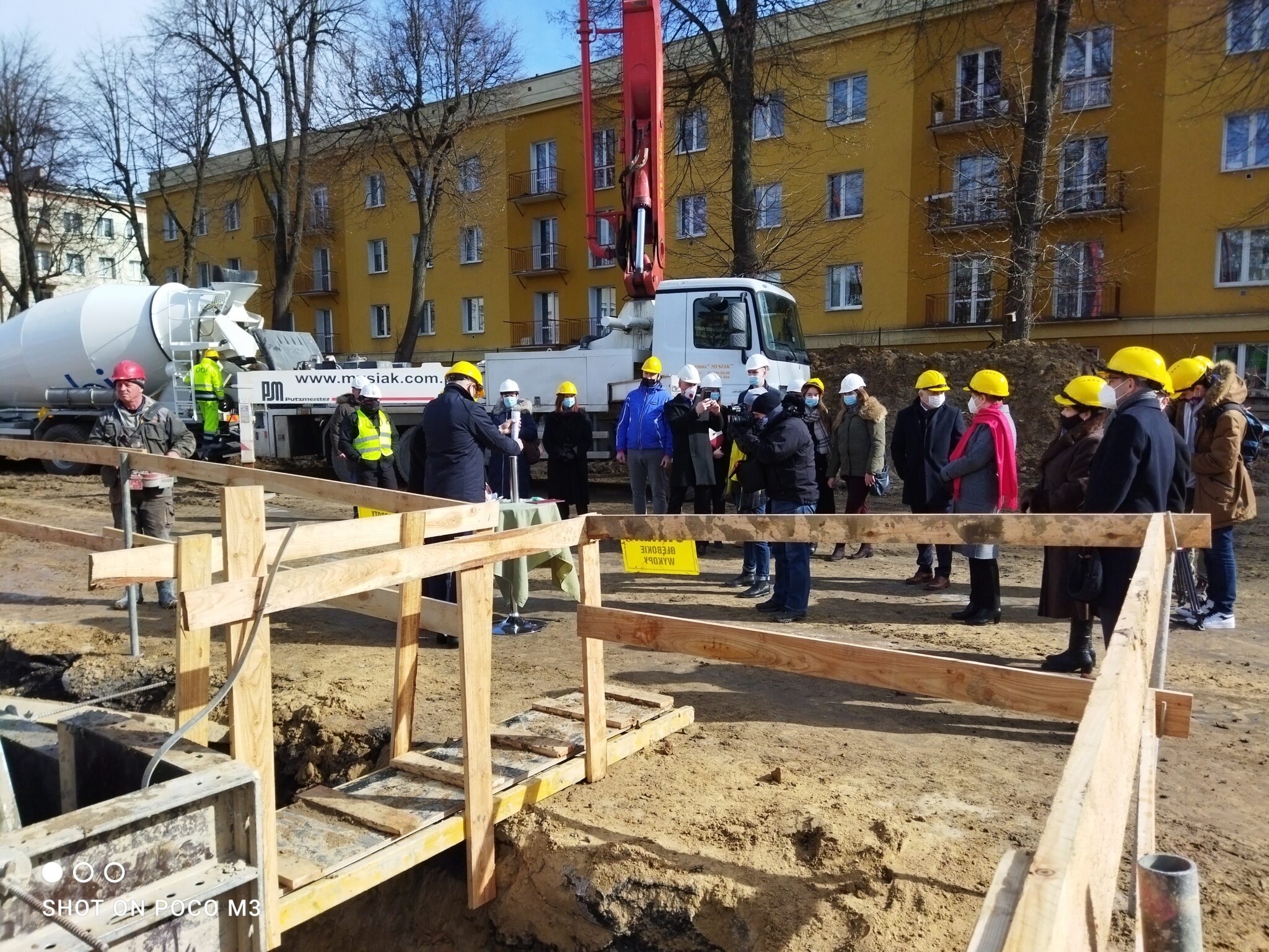 Szerokie ujęcie całej grupy stojącej przy wykopie, z lewej strony widoczna drewniana platforma i zabezpieczenia. Mężczyzna przy stoliku trzyma dokument. W tle betoniarka, ciężarówka i żółty budynek mieszkalny. Grupa składa się z około 20 osób.