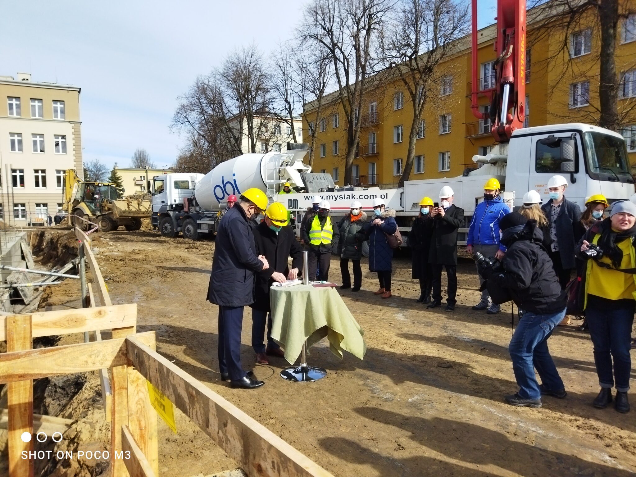 Dwie osoby (prawdopodobnie przedstawiciele władz) stoją przy stoliku z zielonym obrusem, podpisując dokument. W tle – zgromadzeni goście i pracownicy budowy, wykonujący zdjęcia. Widoczna betoniarka z logiem „pbi” oraz koparka z przodu budynku szkolnego.
