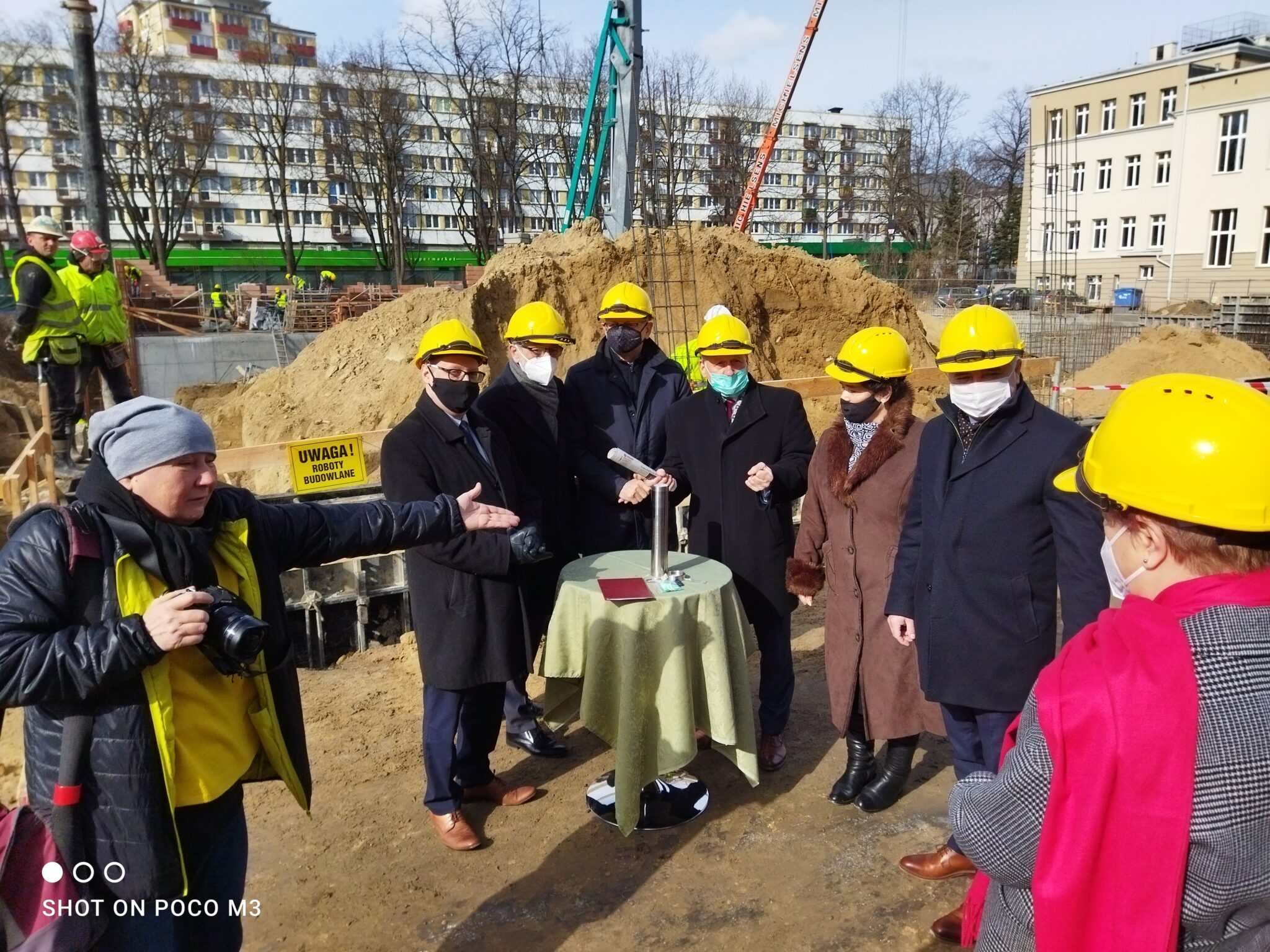 Grupowe zdjęcie – około 10 osób ustawionych wokół stolika z tubą i dokumentami. Wszyscy mają żółte kaski, maseczki, ubrani w zimowe płaszcze. Na pierwszym planie kobieta w czapce i kurtce pokazuje dłonią w kierunku stolika – prawdopodobnie fotografka lub organizatorka wydarzenia.