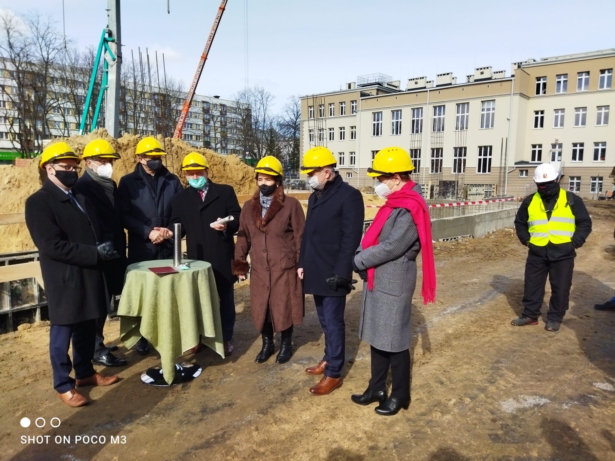 Zdjęcie grupowe – ci sami uczestnicy, co wcześniej, ustawieni wokół zielonego stolika z tubą. W tle góra piachu i rusztowania budowlane, a także budynek szkoły. Po prawej stronie kobieta w szarym płaszczu i różowym szalu, stojąca z założonymi rękami. Mężczyźni i kobiety w eleganckich strojach, gotowi do wmurowania kapsuły czasu.