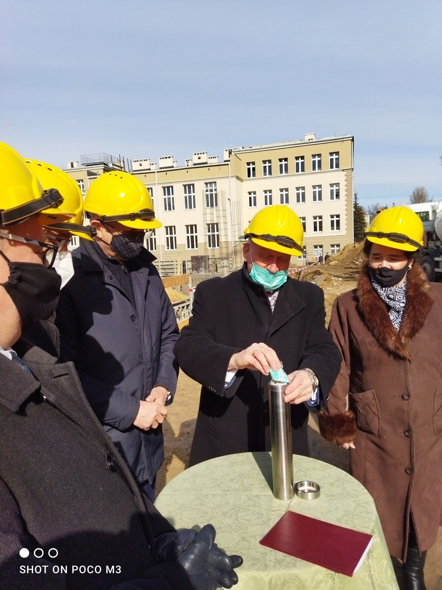 Zdjęcia przedstawiają grupę pięciu osób zgromadzonych przy okrągłym stoliku pokrytym jasnozielonym obrusem. Na środku stołu znajduje się metalowa tuba – tzw. kapsuła czasu – o cylindrycznym kształcie, z otwartym wiekiem obok. Mężczyzna w środku kadru, w czarnym płaszczu i żółtym kasku ochronnym, wkłada do tuby pojedynczą maseczkę ochronną – typową dla pandemii COVID-19. Towarzyszy mu czterech innych uczestników – dwóch mężczyzn i jedna kobieta również w kaskach, płaszczach i maseczkach ochronnych. W tle widoczny jest żółtobiały budynek szkoły.