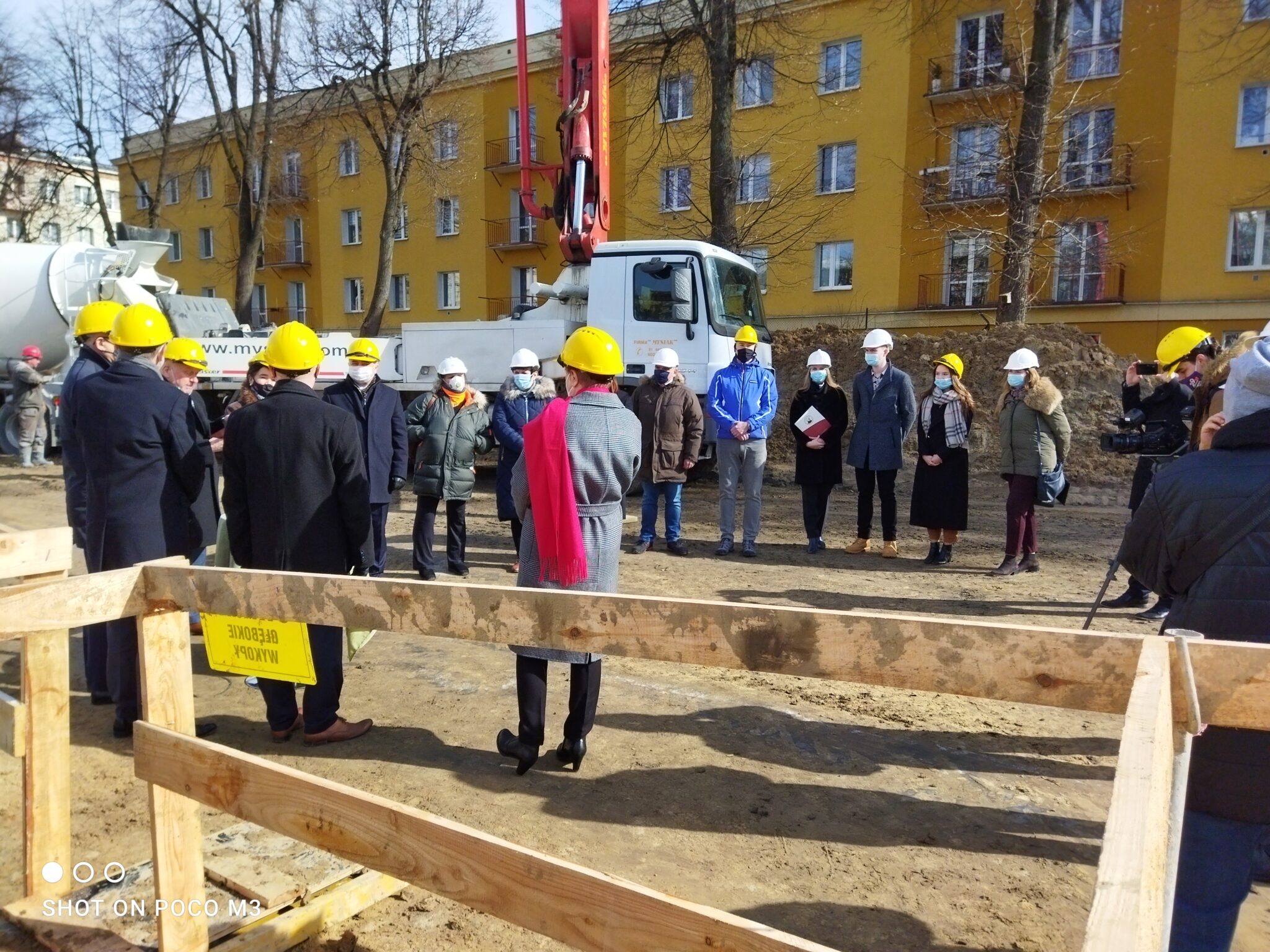 Grupa uczestników uroczystości ustawiona jest w półokręgu przy prowizorycznym zabezpieczeniu wykopu. Po lewej stronie stoi kobieta w szarym płaszczu i czerwonym szalu, z tyłu znajduje się duża biała betoniarka, pojazdy budowlane i żółty blok mieszkalny. Większość osób patrzy w stronę środka półokręgu, gdzie trwa symboliczna część ceremonii.