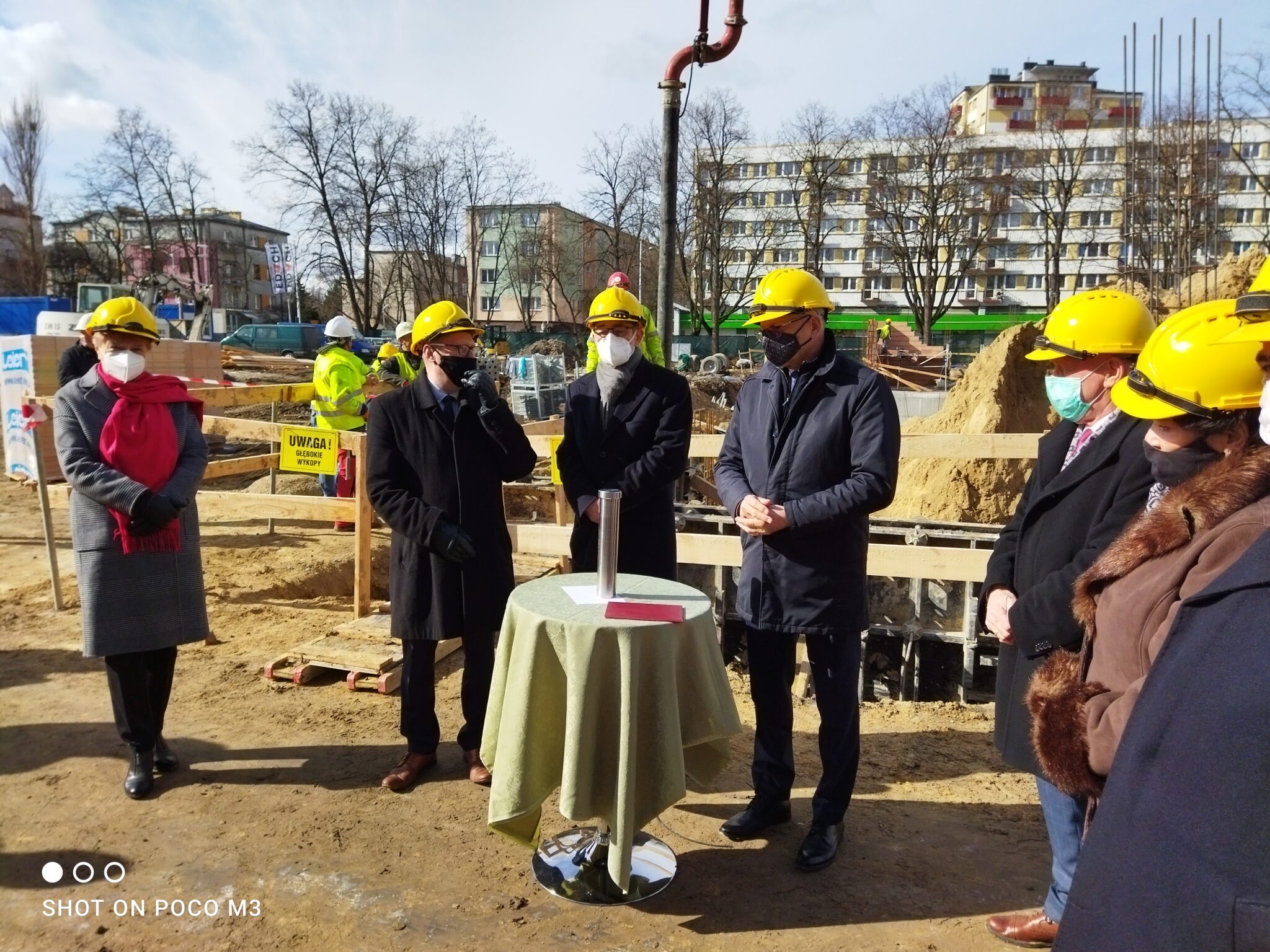 Siedem osób stoi wokół zielonego stolika, na którym znajduje się otwarta kapsuła czasu. Widoczne są składane dokumenty, które prawdopodobnie mają być umieszczone w tubie. Wszyscy uczestnicy ubrani są w eleganckie płaszcze, żółte kaski i maseczki. W tle pracownicy i sprzęt budowlany.