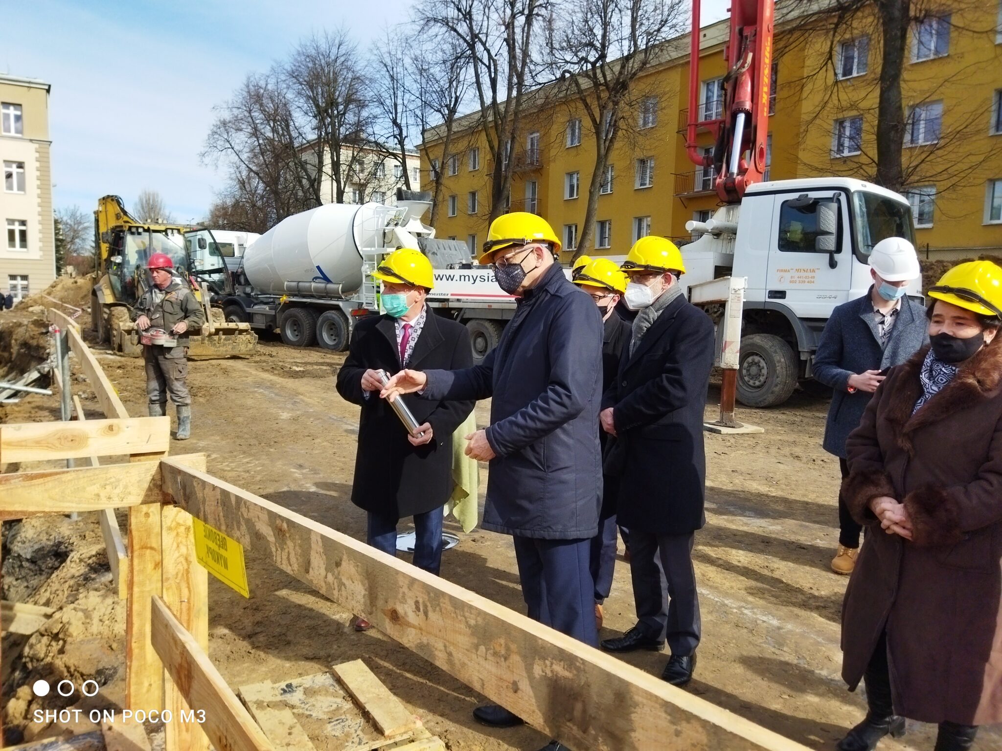 Dwóch mężczyzn w garniturach i żółtych kaskach trzyma metalową tubę – jeden z nich podaje ją drugiemu. Zebrani stoją przy drewnianej konstrukcji wykopu. W tle widać ciężarówkę do betonu i koparkę. Otaczają ich inni uczestnicy uroczystości, w tym kobieta w brązowym futrzanym płaszczu.