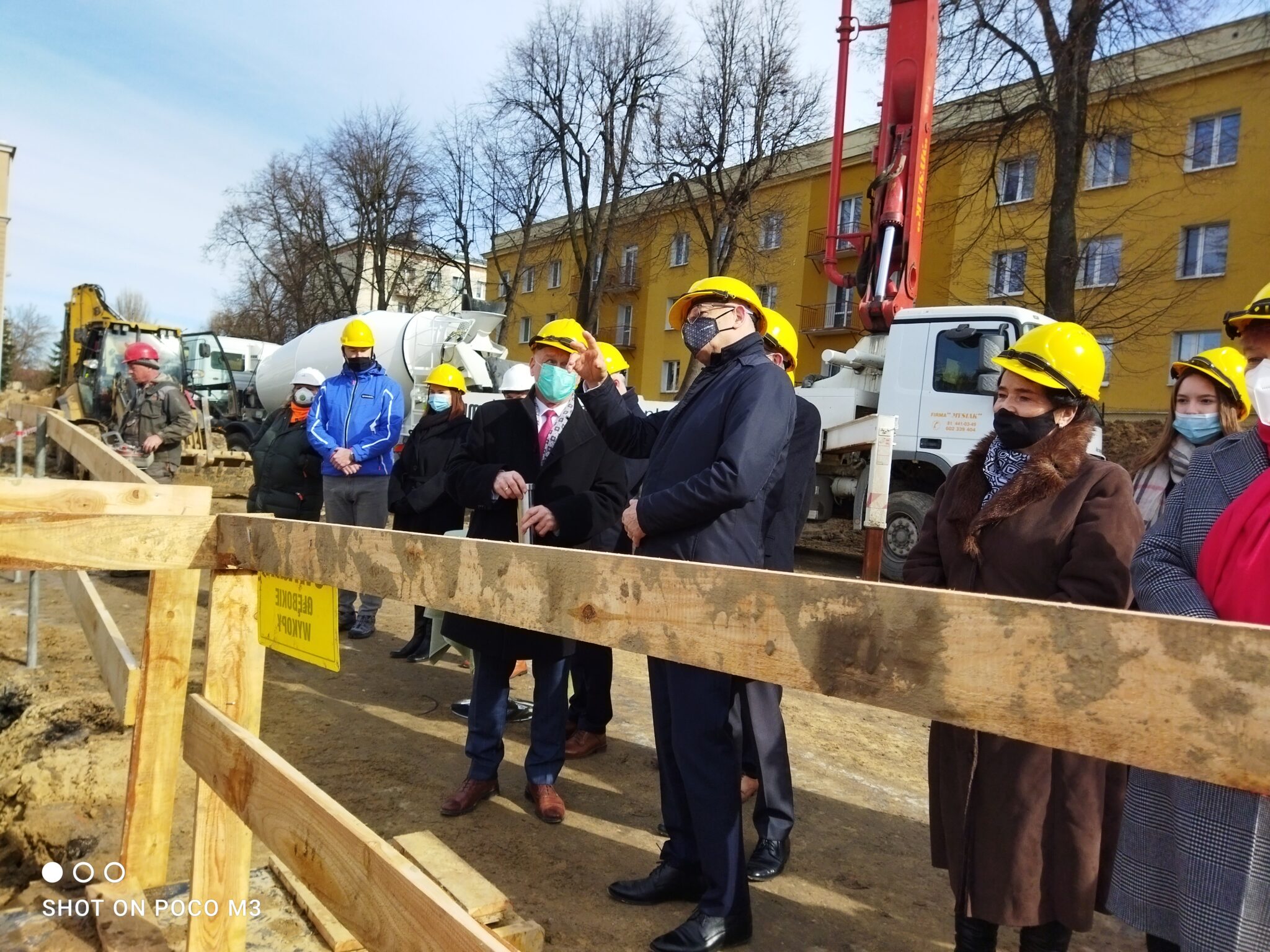 Zdjęcie grupowe z momentu końcowego – osoby ustawione przy wykopie. Mężczyzna w garniturze i kasku pokazuje coś w powietrzu, prawdopodobnie wskazuje przyszłą lokalizację obiektu. Wszyscy uczestnicy uważnie słuchają. W tle sprzęt budowlany i typowa zabudowa mieszkaniowa z blokami.