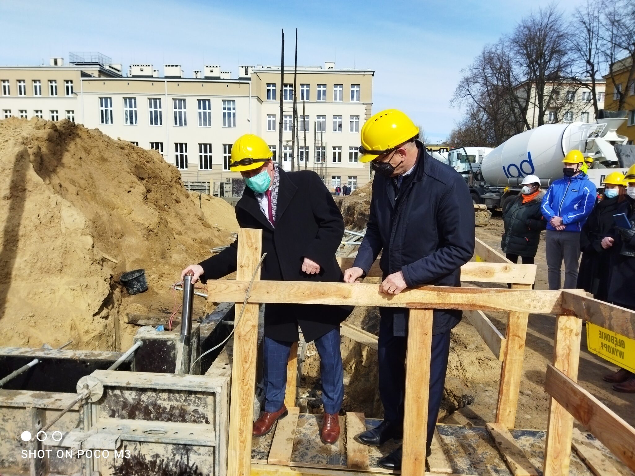 Dwa mężczyźni w garniturach i żółtych kaskach ochronnych stoją na prowizorycznej, drewnianej kładce nad wykopem. Jeden z nich (po lewej) trzyma w ręku stalową tubę – symboliczny kamień węgielny. Obaj obserwują miejsce, do którego tuba ma zostać umieszczona. Kładka zbudowana jest z grubych desek, a całość otoczona jest drewnianą barierką zabezpieczającą. W tle znajduje się wysoka hałda piasku oraz klasyczny, prostokątny budynek szkoły z wieloma oknami.