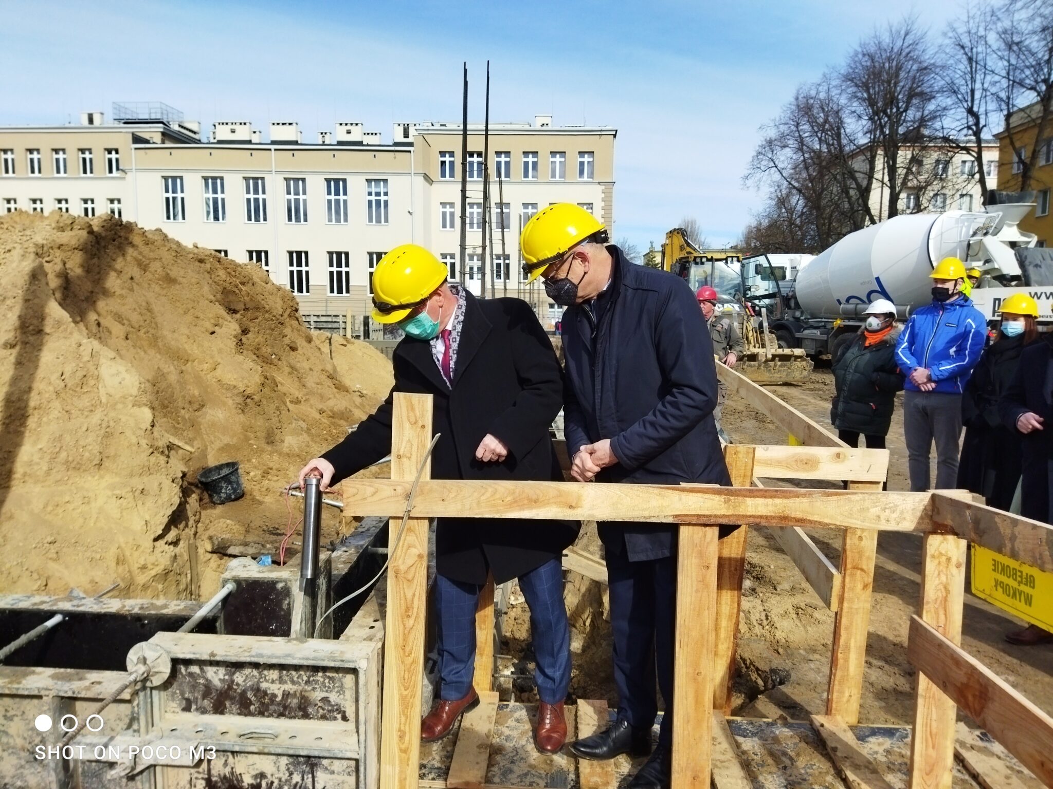 Dwa mężczyźni w garniturach i żółtych kaskach ochronnych stoją na prowizorycznej, drewnianej kładce nad wykopem. Jeden z nich (po lewej) trzyma w ręku stalową tubę – symboliczny kamień węgielny. Obaj obserwują miejsce, do którego tuba ma zostać umieszczona. Kładka zbudowana jest z grubych desek, a całość otoczona jest drewnianą barierką zabezpieczającą. W tle znajduje się wysoka hałda piasku oraz klasyczny, prostokątny budynek szkoły z wieloma oknami.