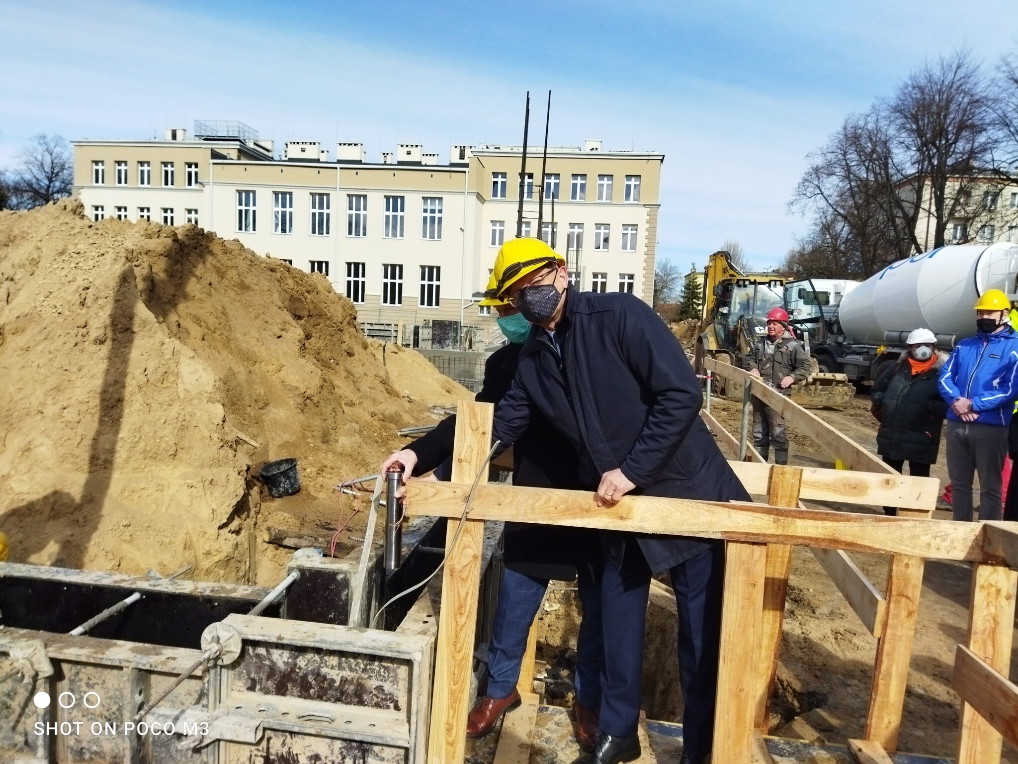 Dwa mężczyźni w garniturach i żółtych kaskach ochronnych stoją na prowizorycznej, drewnianej kładce nad wykopem. Jeden z nich (po lewej) trzyma w ręku stalową tubę – symboliczny kamień węgielny. Obaj obserwują miejsce, do którego tuba ma zostać umieszczona. Kładka zbudowana jest z grubych desek, a całość otoczona jest drewnianą barierką zabezpieczającą. W tle znajduje się wysoka hałda piasku oraz klasyczny, prostokątny budynek szkoły z wieloma oknami.