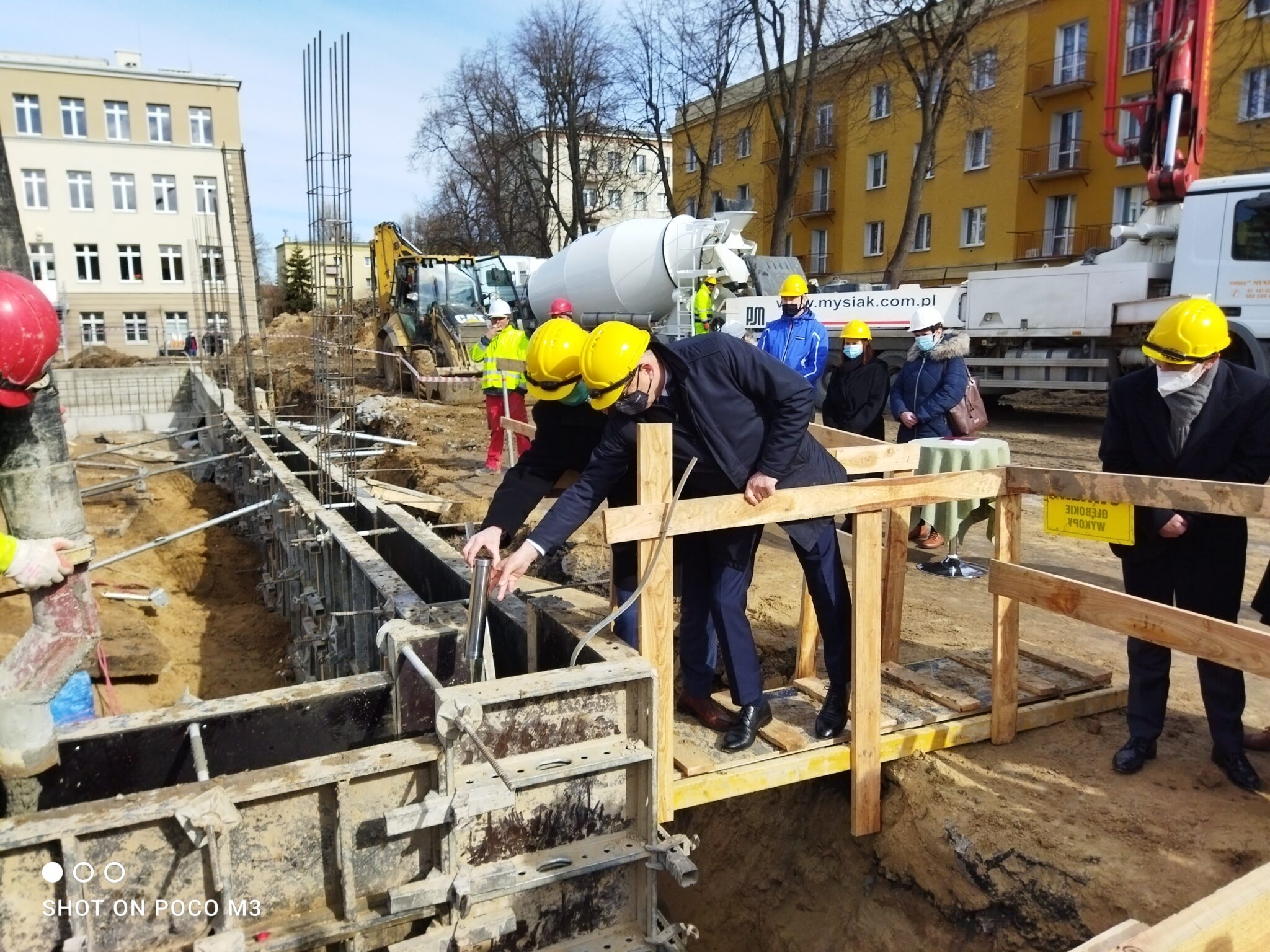 Zdjęcie przedstawia uroczystość wmurowania kamienia węgielnego pod nową inwestycję budowlaną. Scena odbywa się na terenie budowy, w dzień, przy dobrej pogodzie – niebo jest jasne, bezchmurne. Przestrzeń i tło: W tle widoczne są dwa budynki: po lewej stronie klasyczny budynek szkoły lub urzędu, w kolorze beżowym, z dużymi prostokątnymi oknami w równych rzędach. Po prawej stronie widoczny jest żółto-pomarańczowy budynek mieszkalny z balkonami. Za tymi budynkami rosną wysokie, bezlistne drzewa – może to świadczyć o wczesnej wiośnie lub późnej jesieni. Środkowa i przednia część zdjęcia (akcja i osoby): W centrum zdjęcia znajduje się wykop pod fundamenty nowego obiektu, zabezpieczony deskowaniem z metalowymi i drewnianymi elementami. Wykop ma kształt prostokątny i sięga na głębokość około 1,5 metra. Na pierwszym planie, przy krawędzi wykopu, zbudowano prowizoryczną drewnianą platformę z poręczami z desek, służącą jako miejsce symbolicznego wmurowania kamienia węgielnego. Na platformie stoją dwaj mężczyźni w garniturach i żółtych kaskach ochronnych – obaj pochylają się, jeden z nich (bardziej z przodu) trzyma metalową tubę – symboliczny „kamień węgielny”, który właśnie wkłada do wcześniej przygotowanego otworu w deskowaniu fundamentów. Obaj mężczyźni mają na sobie maseczki ochronne, co może sugerować, że zdjęcie wykonano w czasie pandemii COVID-19. W tle za platformą stoją osoby w odblaskowych kamizelkach i kaskach – prawdopodobnie pracownicy budowy. Widoczny jest również betoniarka (biała ciężarówka z dużym bębnem do mieszania betonu) oraz koparka z podniesionym ramieniem. Po prawej stronie, za platformą, stoi grupa osób – są to najprawdopodobniej zaproszeni goście. Wszyscy są ubrani w zimowe kurtki, mają maseczki i kaski ochronne. Wśród nich znajdują się dwie kobiety i kilku mężczyzn. Na drewnianej barierce platformy widnieje żółta tabliczka z napisem „UWAGA! WYKOP GŁĘBOKI”. Inne szczegóły: – W lewym dolnym rogu zdjęcia znajduje się znak wodny z napisem „SHOT ON POCO M3” – to informacja o modelu telefonu, którym wykonano zdjęcie. – Na betoniarce widnieje adres strony internetowej: „www.mysiak.com.pl” – prawdopodobnie to firma odpowiedzialna za inwestycję budowlaną. Symbolika i kontekst: Czynność wmurowania tuby z dokumentami do fundamentu symbolizuje rozpoczęcie budowy lub ważnego etapu inwestycji, często z udziałem władz lokalnych, inwestorów lub mediów. Może to być szkoła, budynek użyteczności publicznej lub inny obiekt o znaczeniu społecznym.