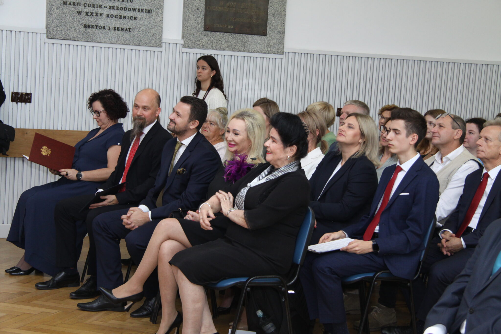 W pierwszym rzędzie zasiadają dorośli w eleganckich strojach — garnitury, sukienki, apaszki. Niektórzy z nich trzymają dokumenty lub teczki. Na twarzach skupienie i zaangażowanie. W tle widać marmurowe tablice i panele ścienne w jasnym kolorze.