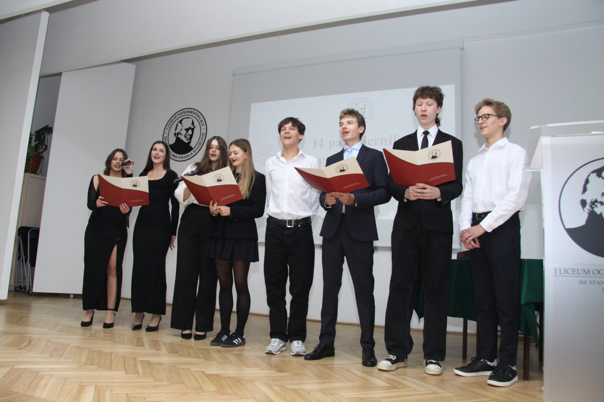 Grupa elegancko ubranych uczniów stoi na scenie. Czworo z nich trzyma w dłoniach teczki brązowo-kremowe. W tle logo szkoły. Młodzież ubrana na galowo