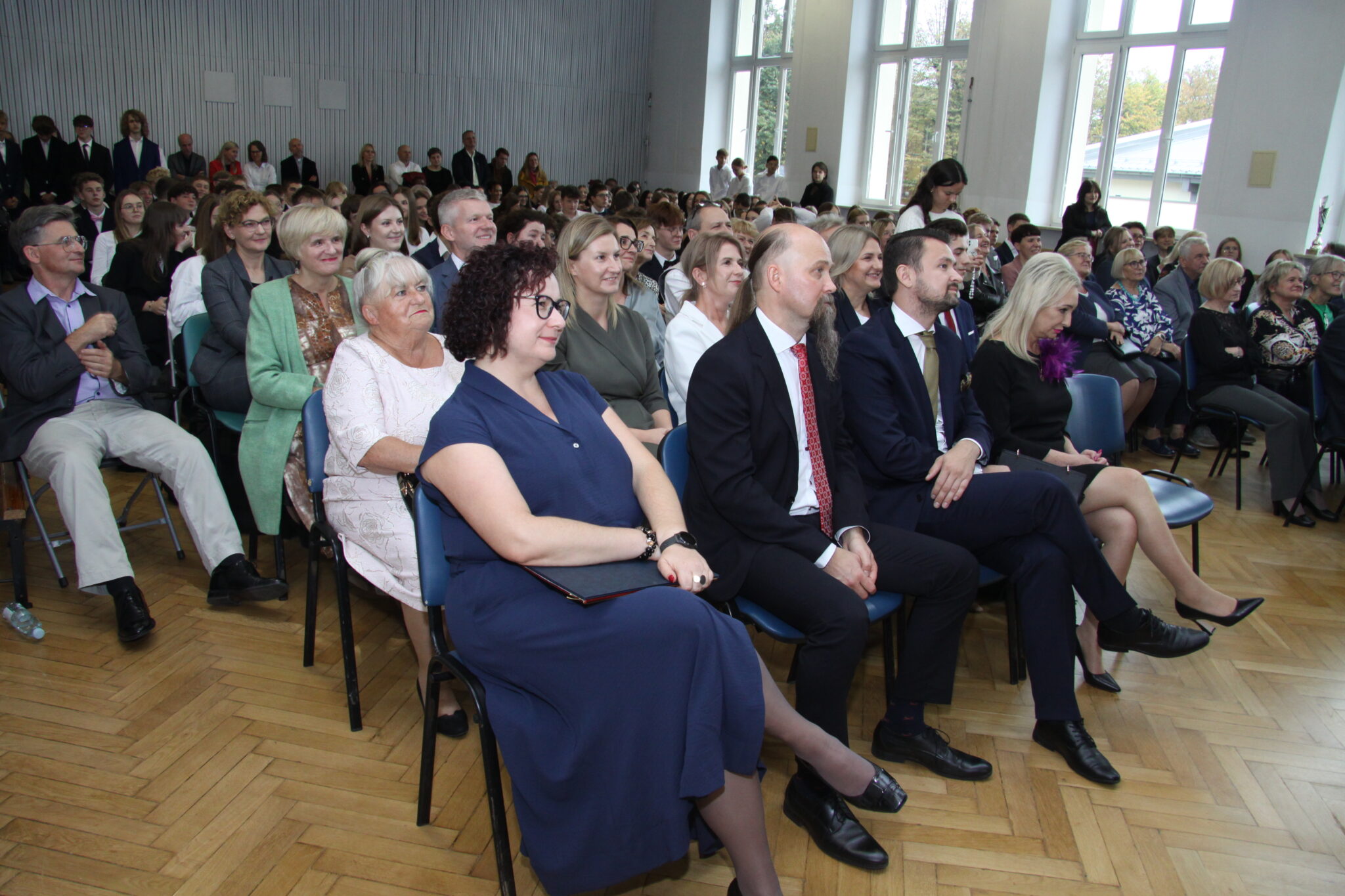 Dziesiątki gości siedzą na krzesłach podczas uroczystości w auli szkolnej. Na pierwszym planie konieta w niebieskiej sukience o czarnych kręconych włodach i mężczyzna z siwą brodą w garniturze. W tle 3 okna.
