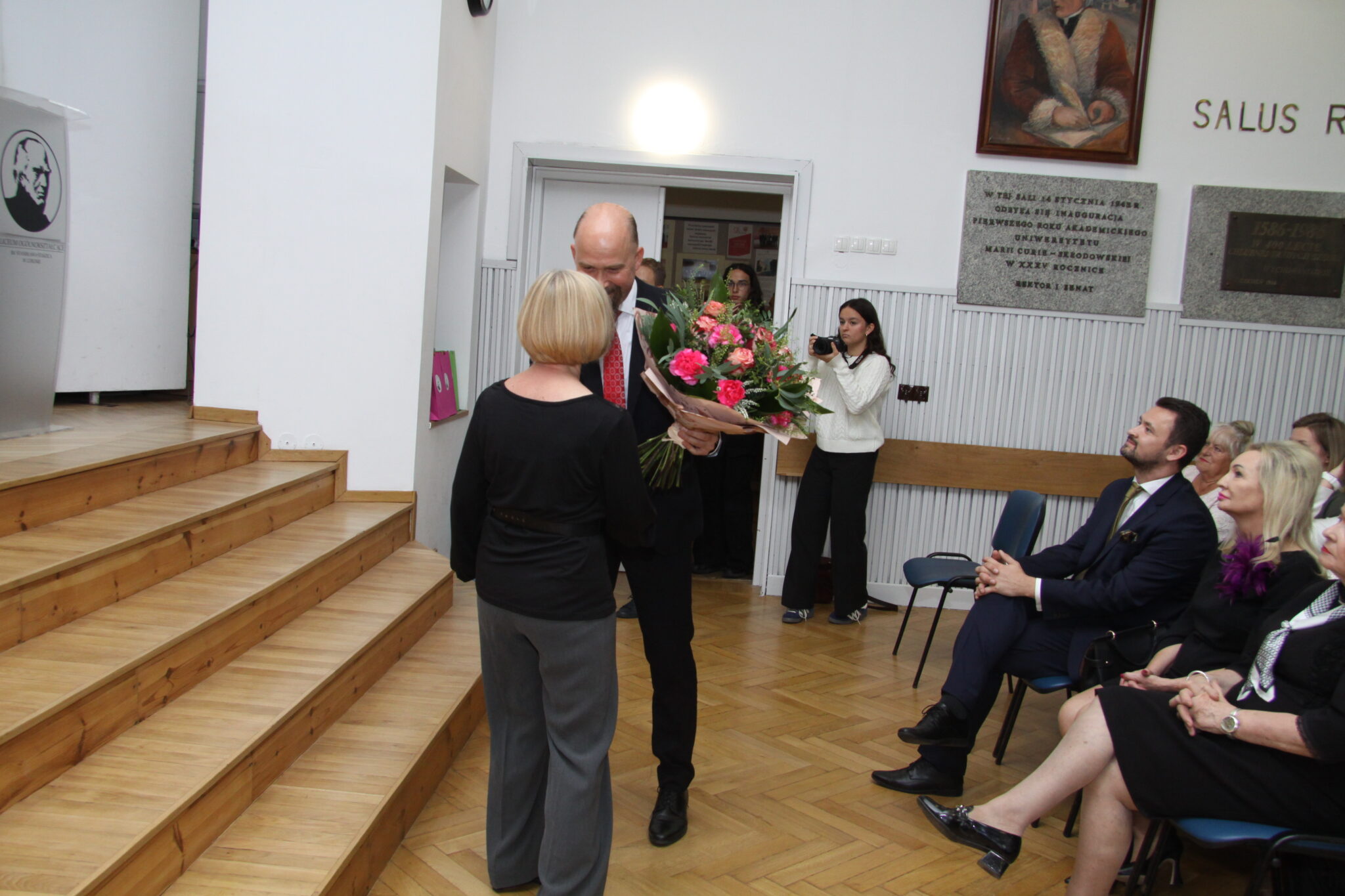 Mężczyzna z siwą brodą wręcza bukiet kwiatów kobiecie i krótkich blond włosach. Scena z auli szkolnej podczas uroczystości. Mężczyzna jest w garniturze ciemnym, kobieta w ciemnym sweterku i szarych spodniach.