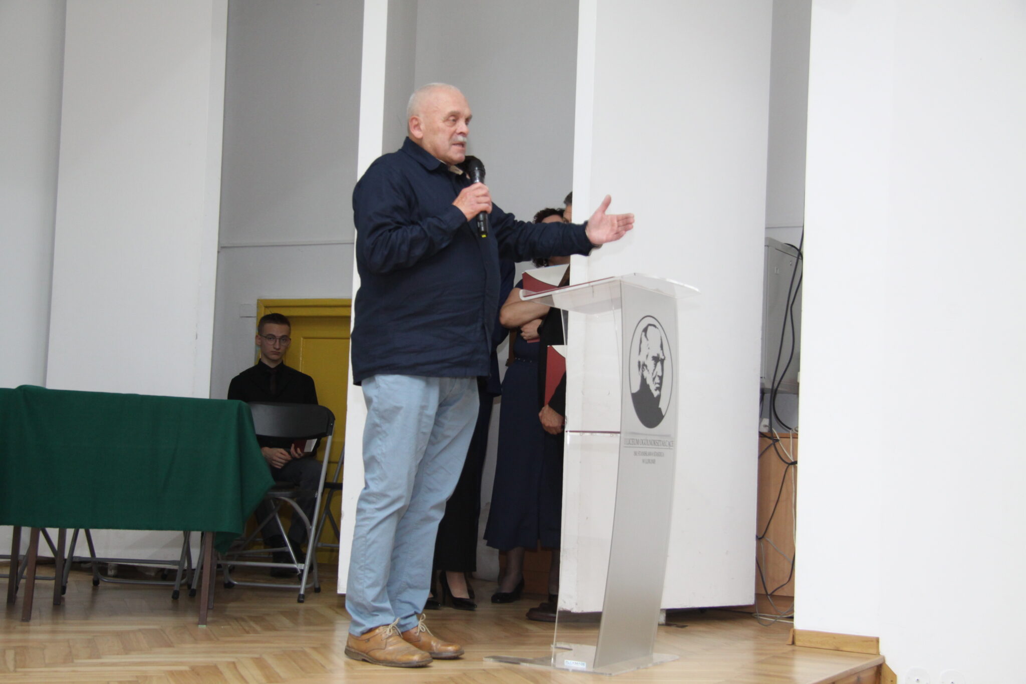 Starszy mężczyzna o słusznej posturze przemawia z mikrofonem w ręku na scenie w auli szkolnej. Jest ubrany w ciemną koszulę i dżinsowe spodnie. Stoi przy mównicy i gestykuluje ręką.